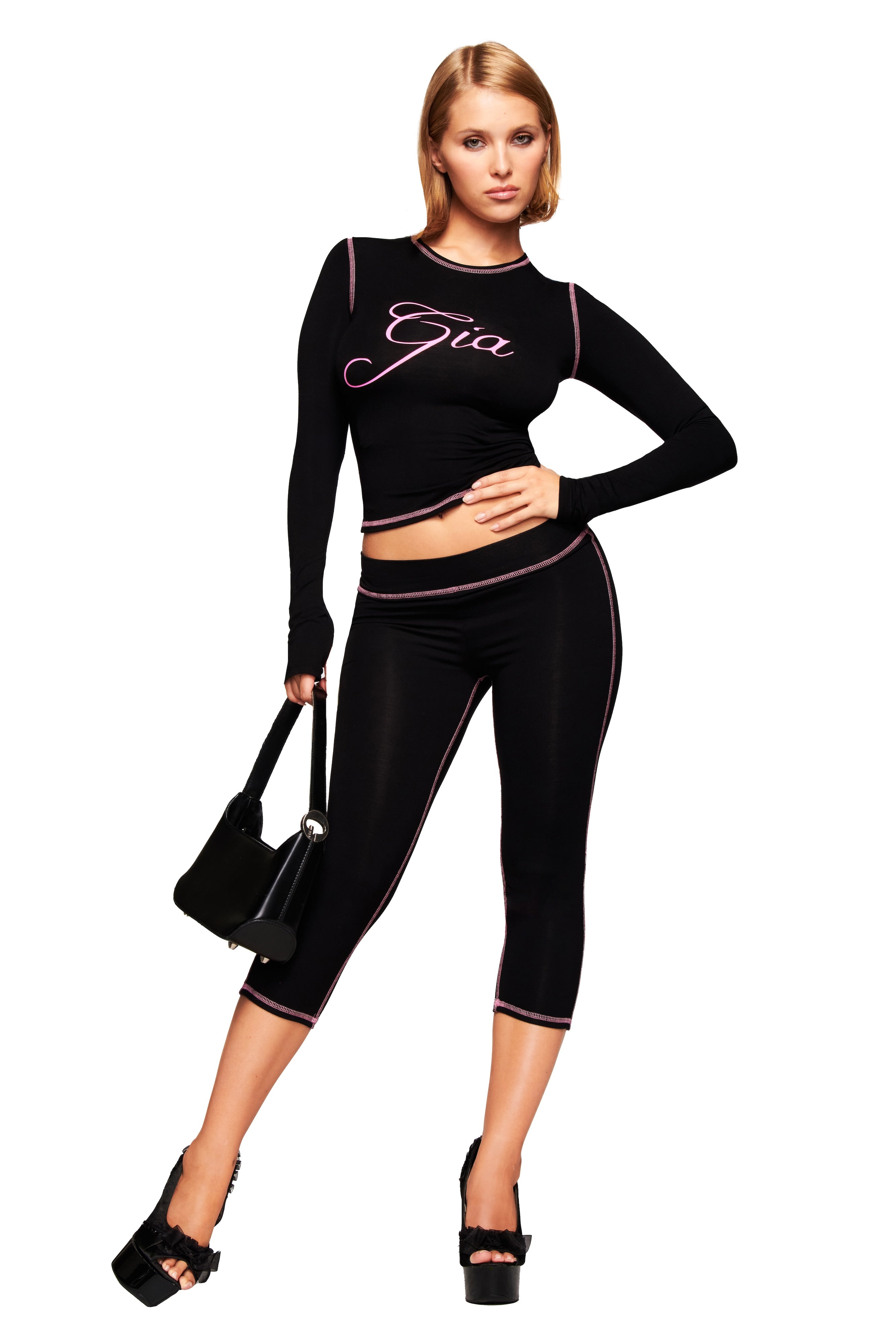 A woman poses confidently in an I.AM.GIA BLARE CAPRI - BLACK set with matching black long-sleeve crop top, platform heels, and a black handbag against a white background.