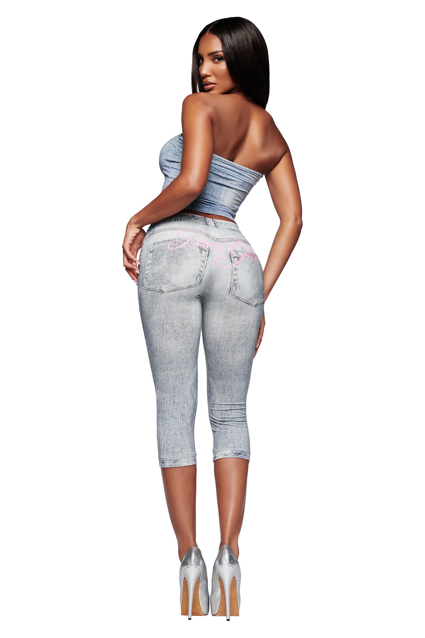 A woman with long straight hair models the I.AM.GIA BLARE CAPRI - DENIM TROMPE L'OEIL pant and a strapless denim top, posing with her back to the camera in silver high heels and looking over her shoulder.