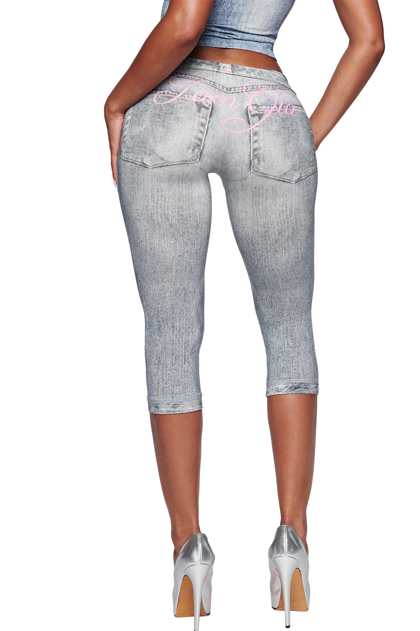 A woman is shown from the back in I.AM.GIA’s BLARE CAPRI - DENIM TROMPE L'OEIL, light gray capri pants with a faux-denim look, two back pockets, pink script on the waistband, and paired with silver high heels.