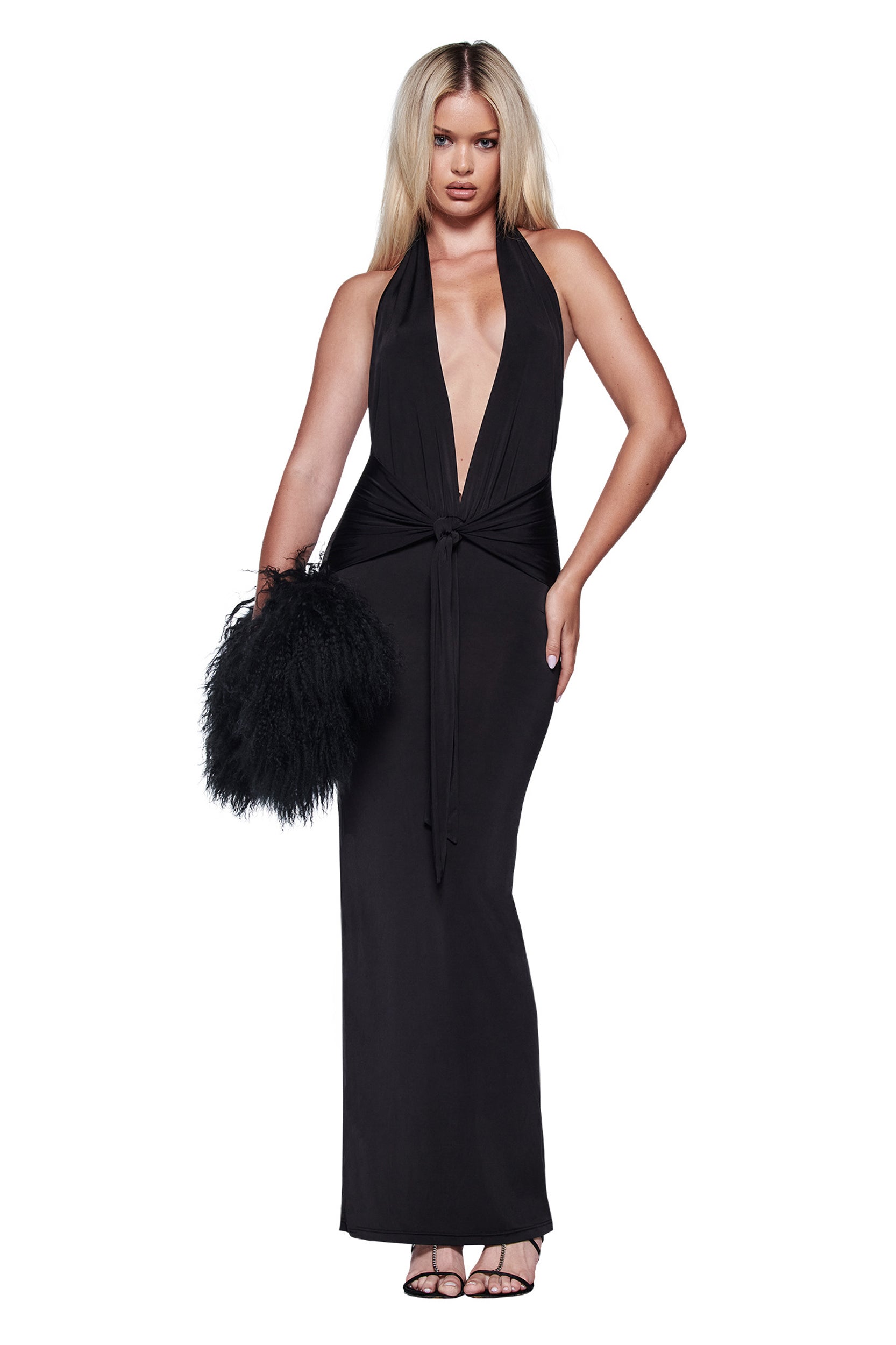 A woman with long blonde hair models the I.AM.GIA ZIVA PLUNGE TIE MAXI DRESS in black, featuring a plunging V neckline and front tie. She holds a black feathered clutch and heels, posing against a white background.