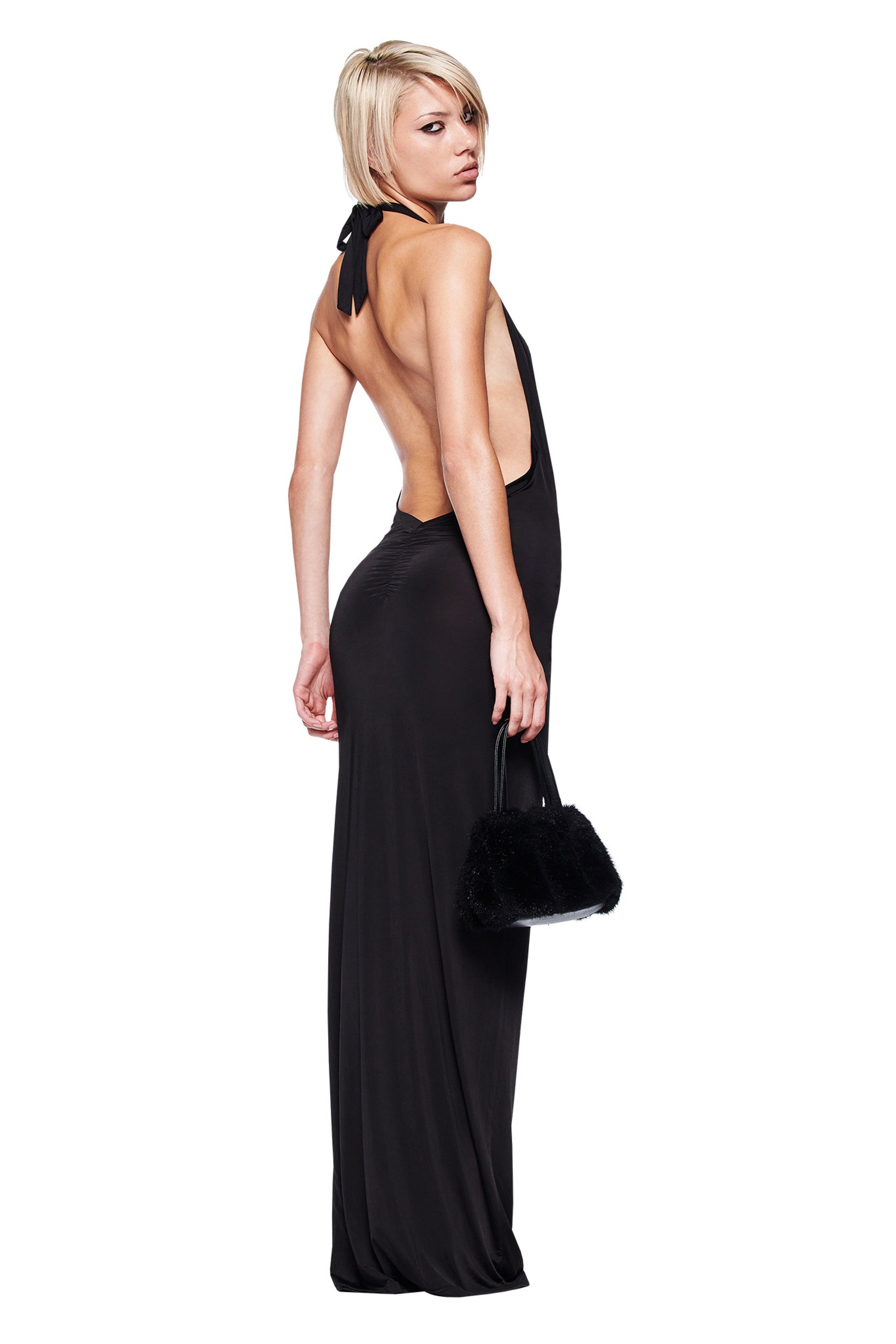 A woman with short blonde hair models the I.AM.GIA ZIVA Plunge Tie Maxi Dress in black, featuring an open back. She stands sideways, looking over her shoulder, and holds a small black handbag against a white background.
