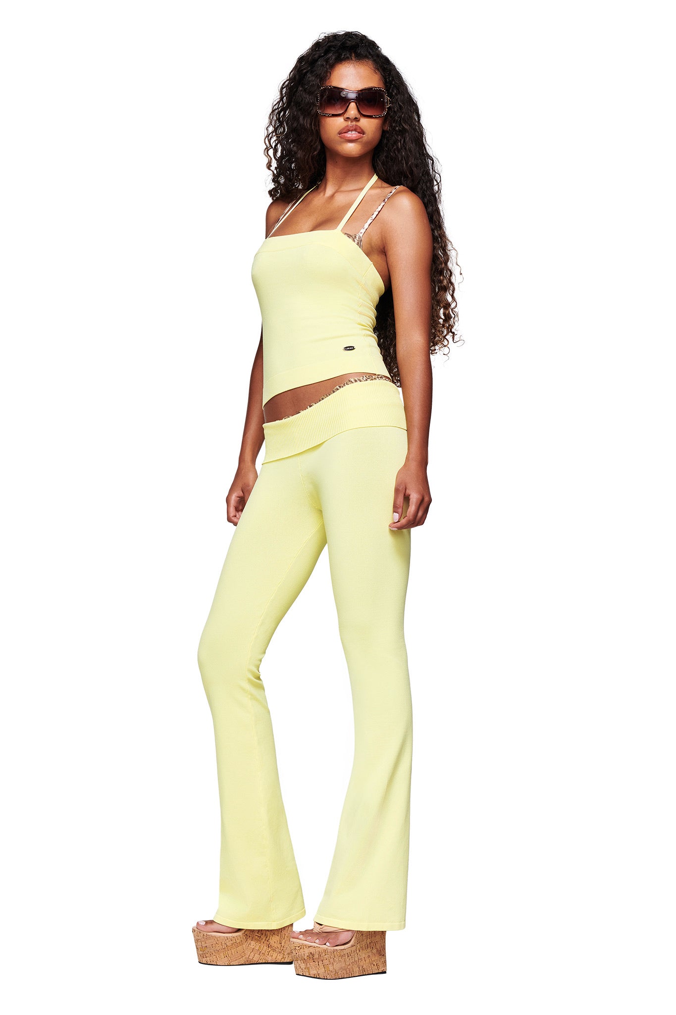 A woman with long curly hair wears sunglasses, a light yellow spaghetti strap crop top, I.AM.GIA's MORGANA 2.0 PANT in Baby Yellow, and platform sandals, standing against a white background.