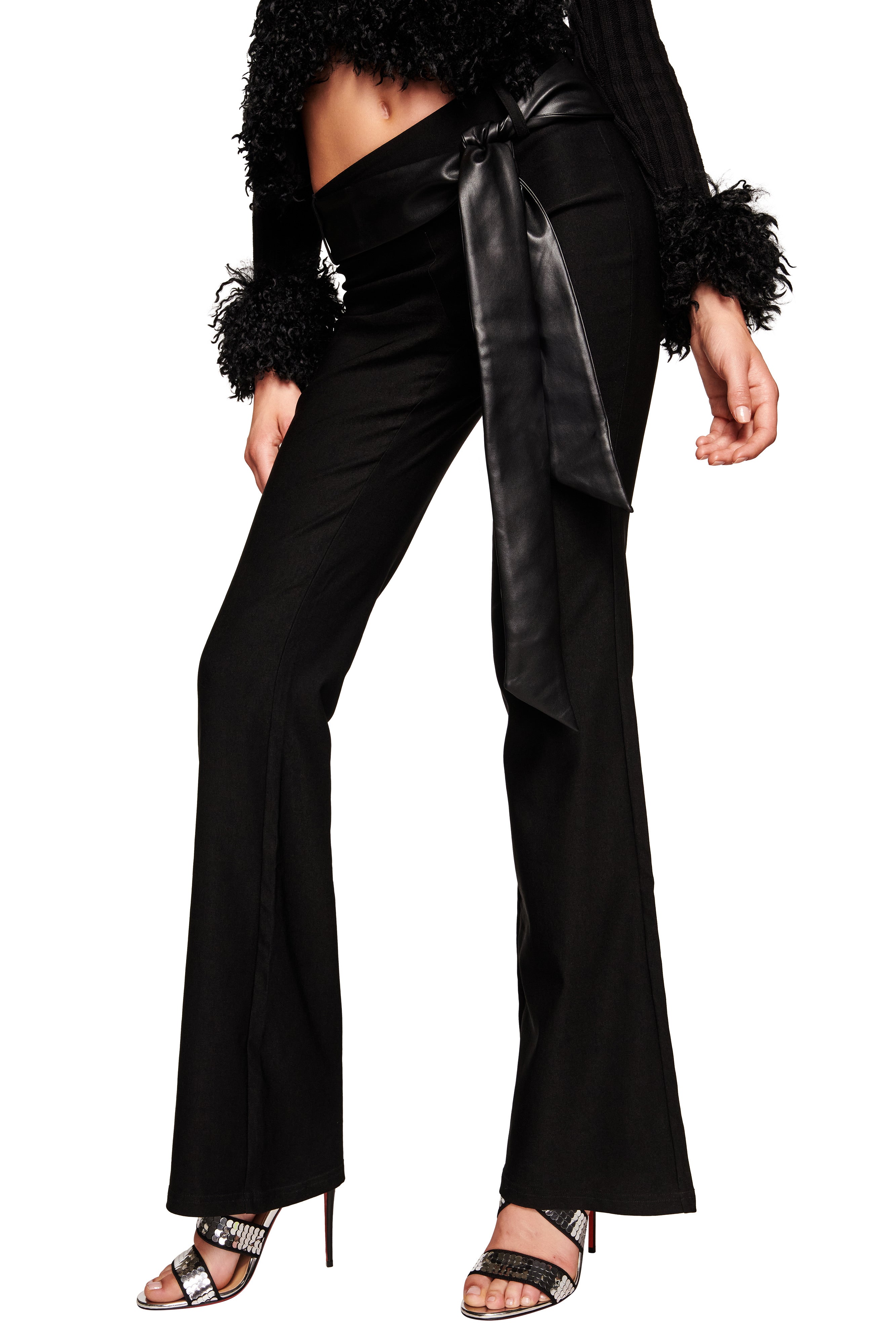 Only the lower body and torso are visible, showing a person wearing I.AM.GIA's FELIZ PANT - BLACK with a wide faux leather belt, a black textured long-sleeve top with furry cuffs, and black high-heeled sandals.