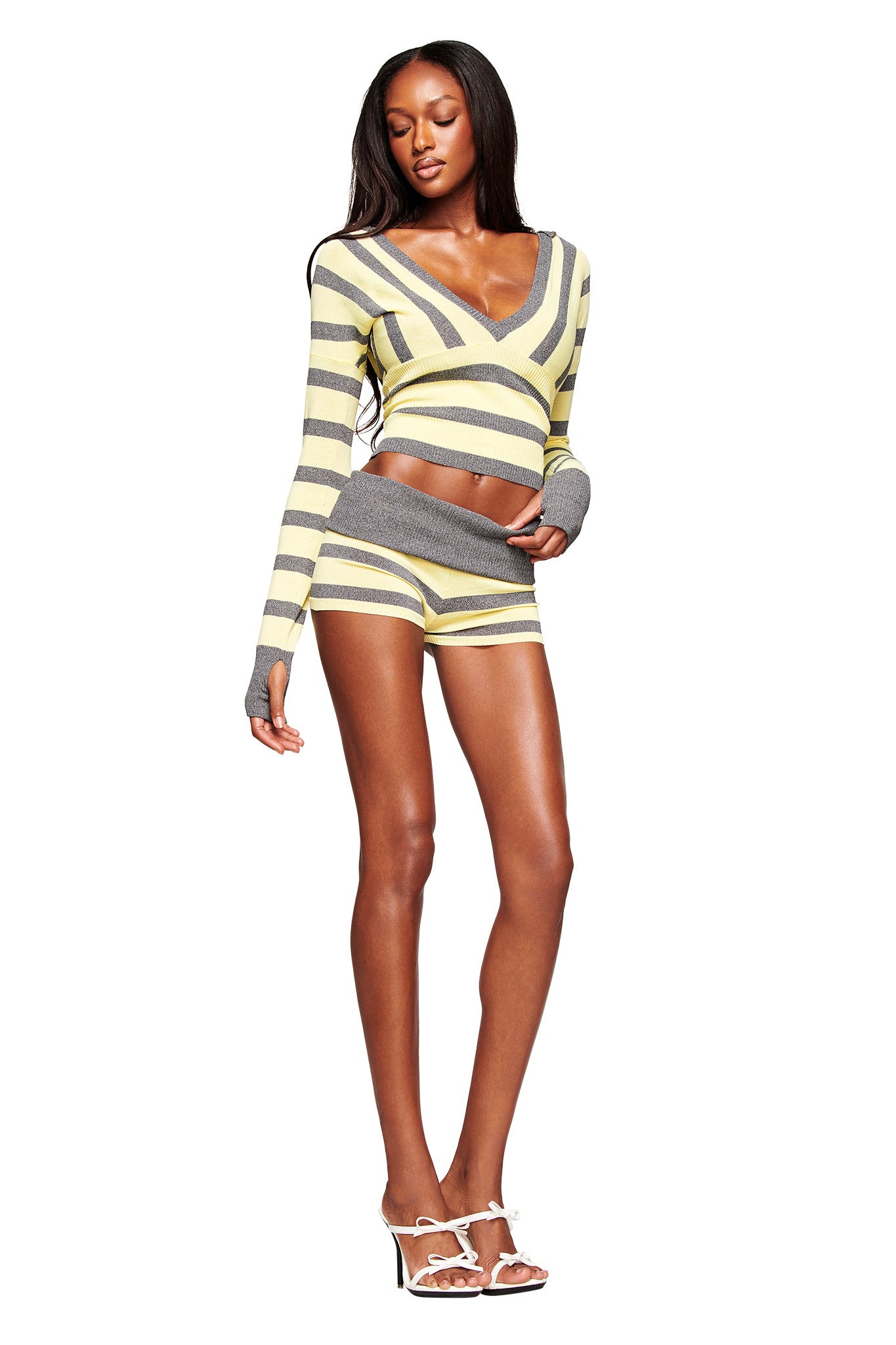 A woman poses confidently in I.AM.GIA's SUZI SHORT - YELLOW AND GREY STRIPE, styled with an adjustable waistband and paired with a long-sleeve crop top and white high-heeled sandals against a plain white background.