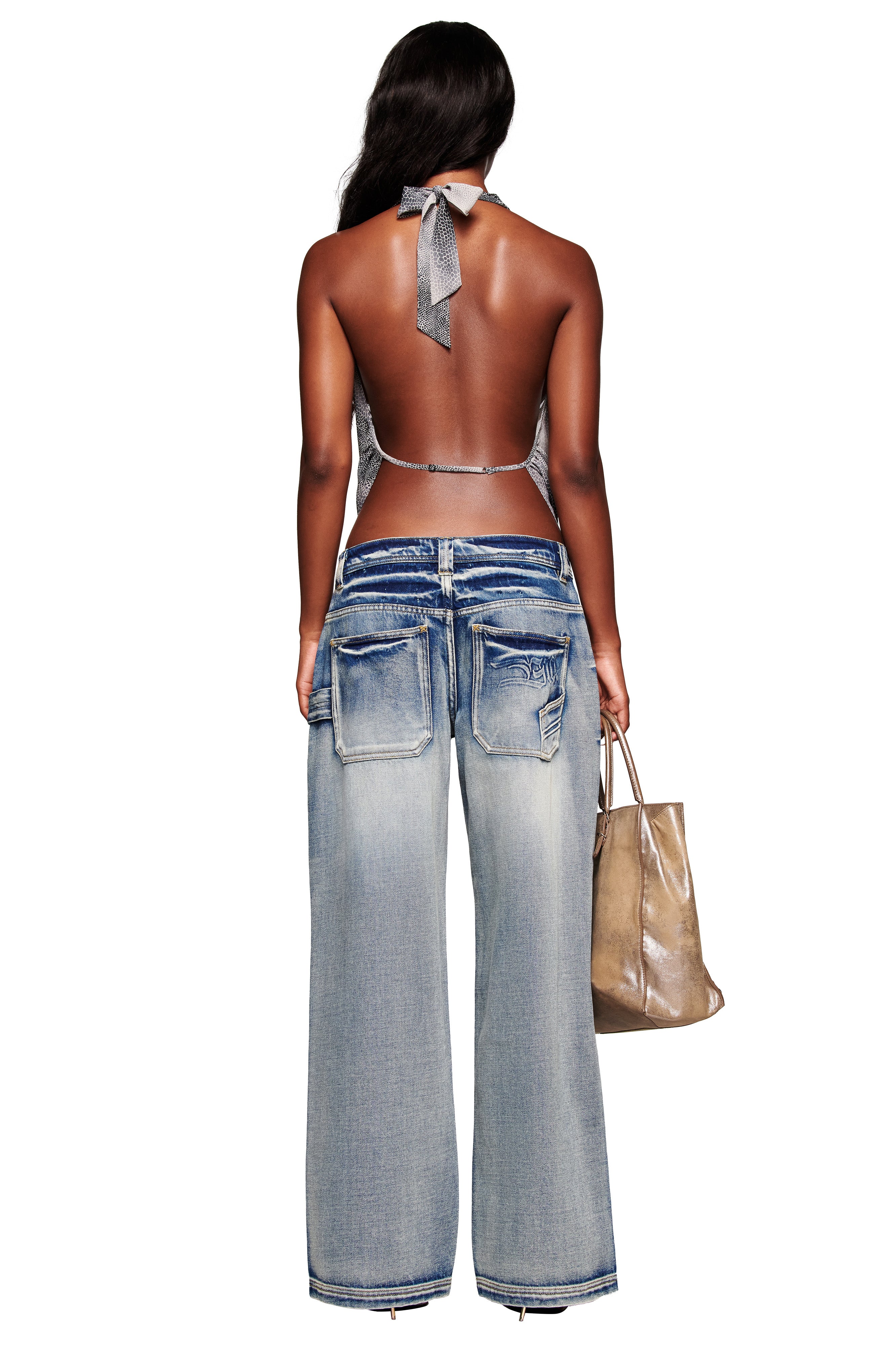A woman with long dark hair is shown from behind wearing I.AM.GIA's PALISADE JEAN - BLUE, a baggy light-wash style with large back pockets, paired with a halter top and holding a metallic tote bag against a white background.