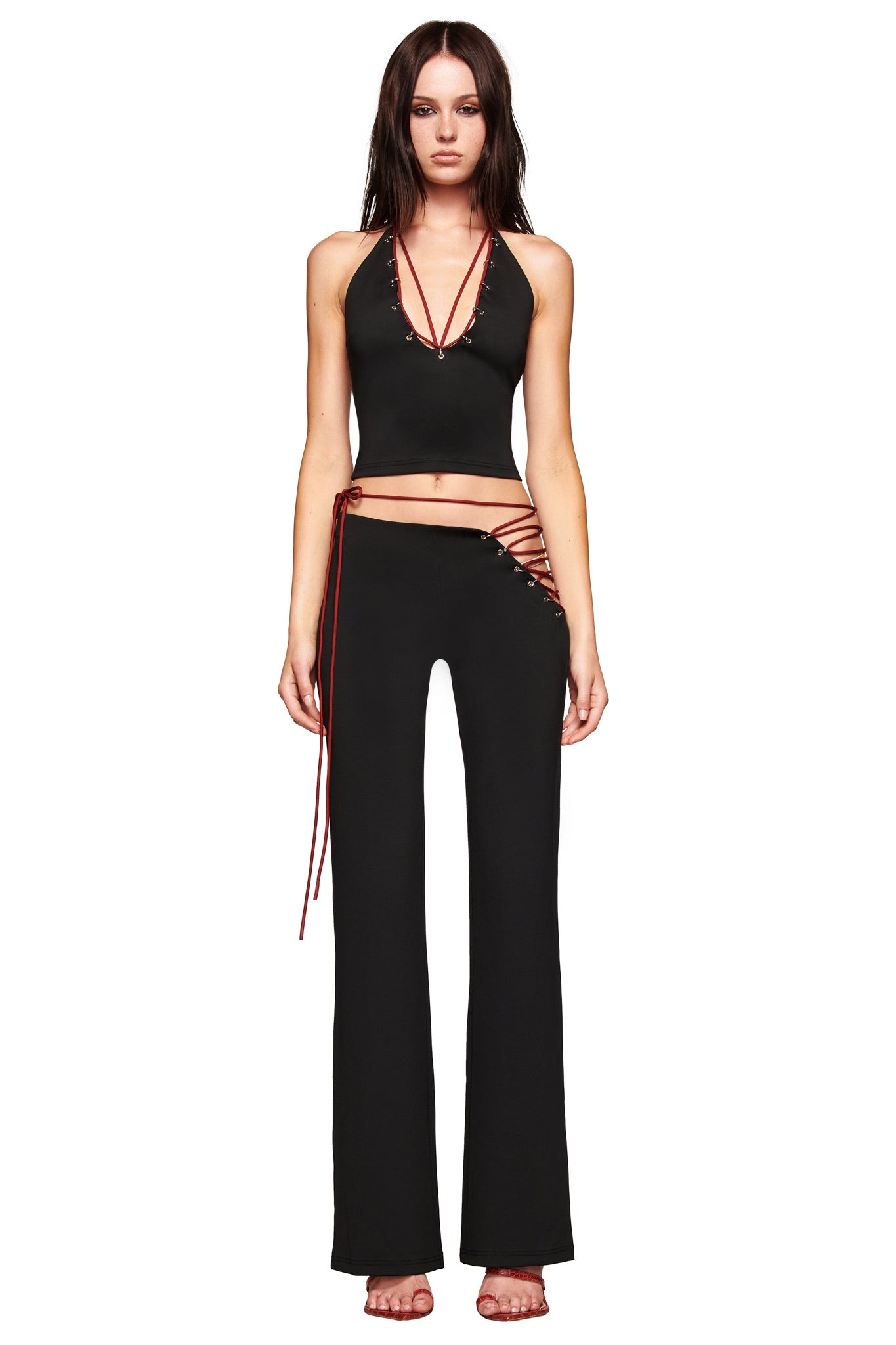 A woman stands facing forward in I.AM.GIA's ONYX PANT - BLACK, a black eyelet pant with red lace-up accents and a low-rise fit, styled with a black halter crop top with red string details and open-toe heels against a white background.