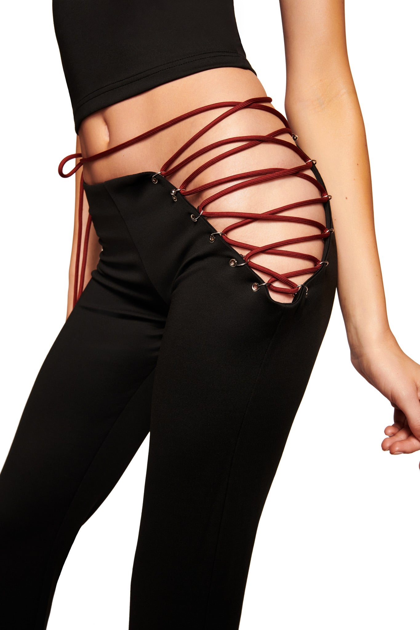 A person wears the I.AM.GIA ONYX PANT - BLACK, a low-rise black pant with exposed hips, featuring a crisscross of red laces through metal eyelets, styled with a black top.