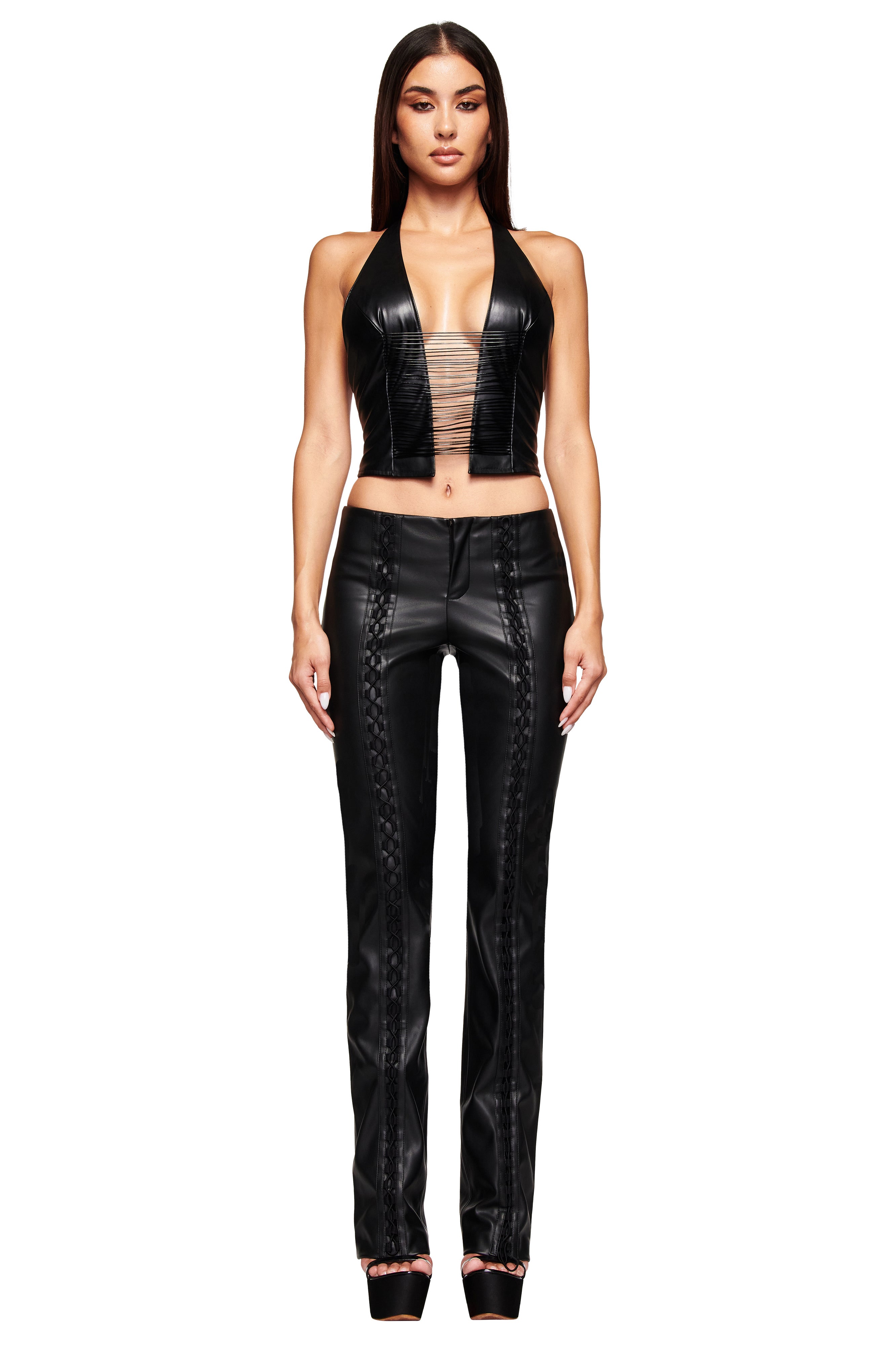 A woman faces forward wearing the ALTAIR TOP - BLACK by I.AM.GIA, featuring waxed cord straps, paired with a sheer nude bandeau, black stitched leather pants, and black platform heels.