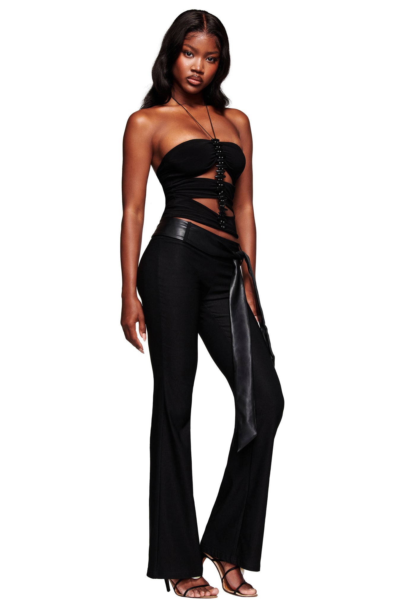 A woman stands confidently in the I.AM.GIA FENNICK TOP - BLACK, paired with black flared pants and high-heeled sandals. She has long, wavy hair and a neutral expression.