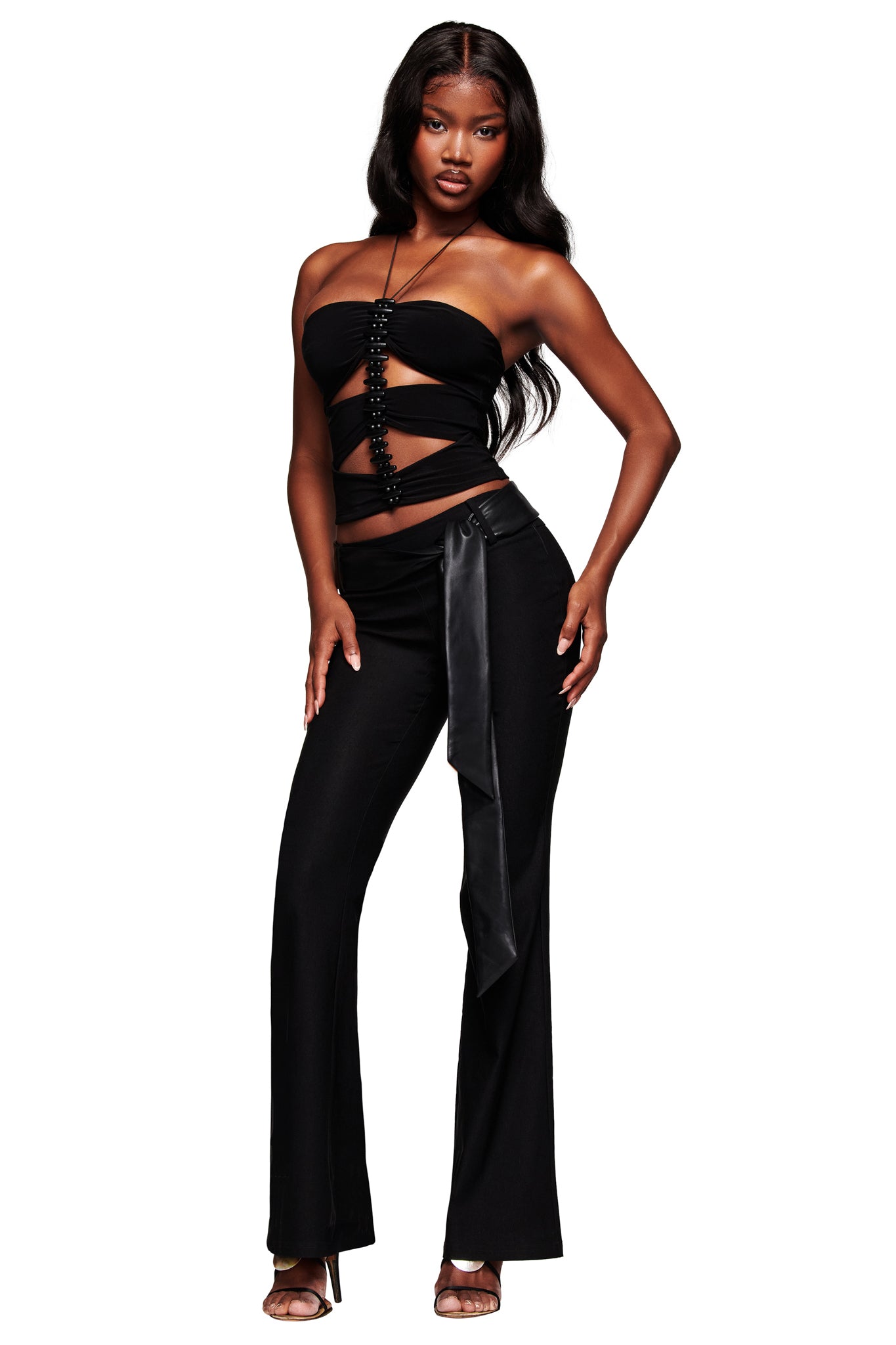 A woman poses confidently in the I.AM.GIA FENNICK TOP - BLACK, a strapless cut-out top with beaded details, styled with high-waisted pants, a satin sash, and heeled sandals.