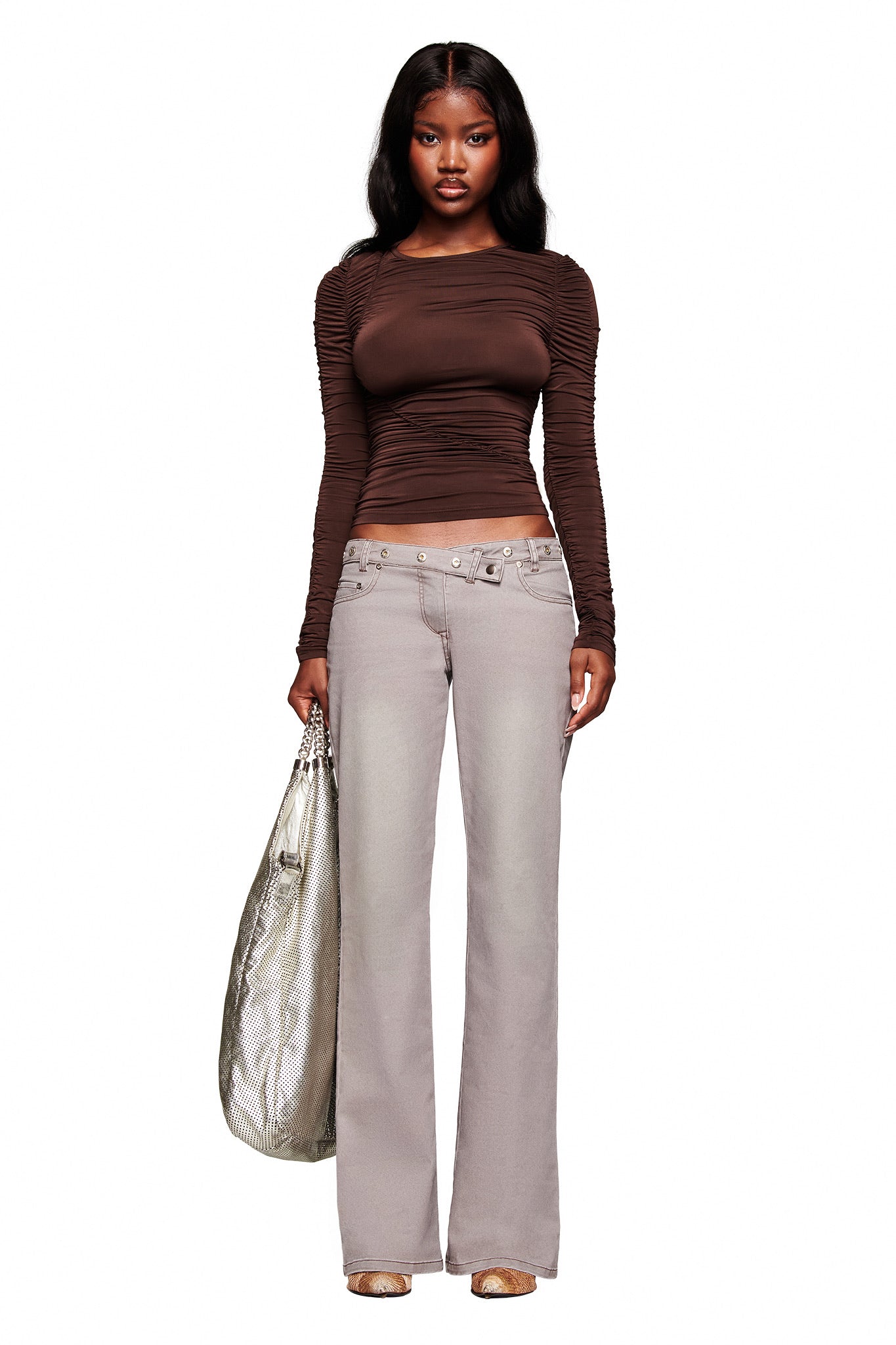 A woman faces forward against a white background, wearing the I.AM.GIA GREER PANT in grey with a fitted long-sleeve brown top, neutral shoes, a large silver bag, and long wavy hair.