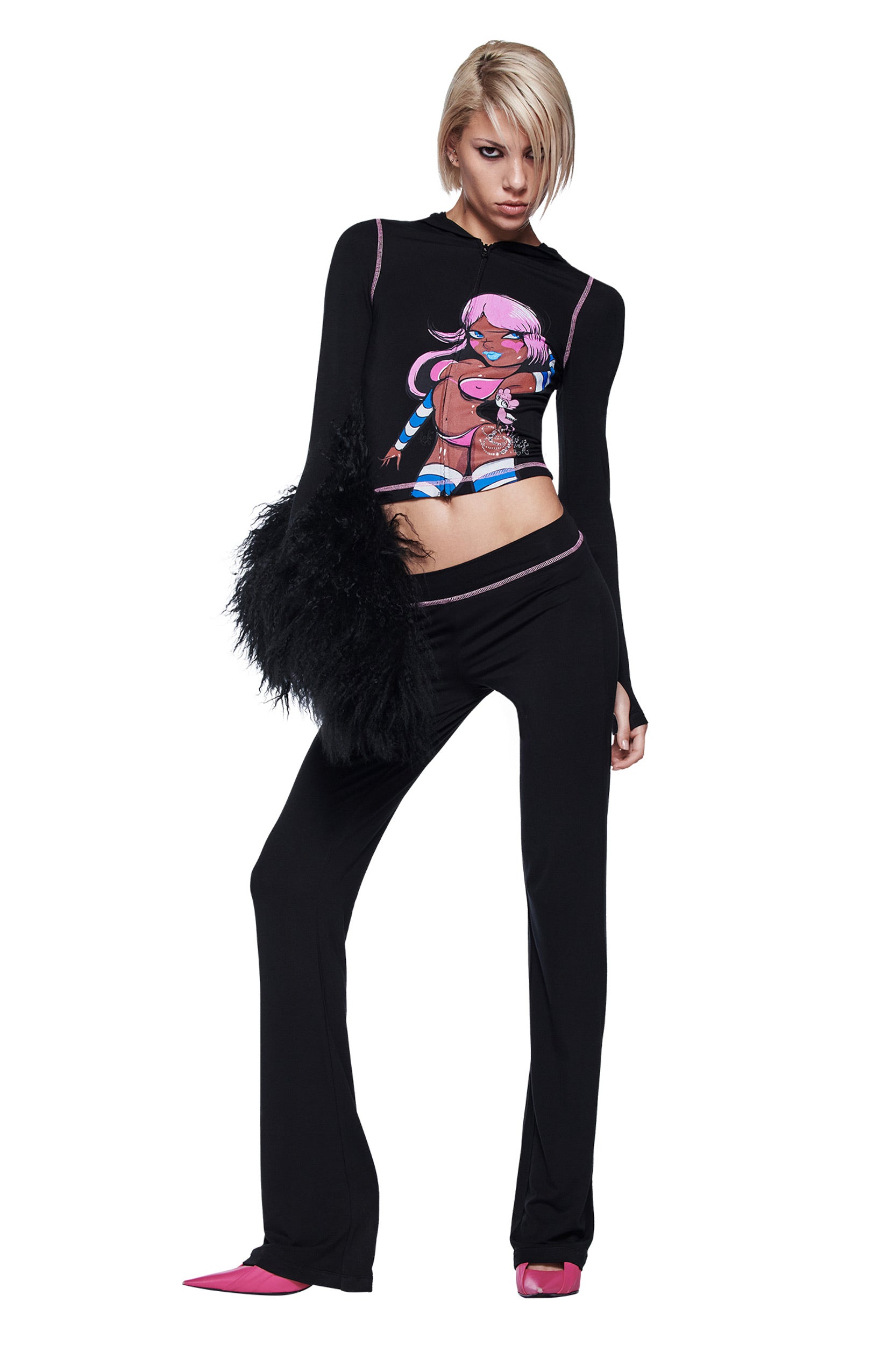 A person with short blonde hair wears a black I.AM.GIA long-sleeve crop top featuring a cartoon, FAFI BLARE PANT - BLACK by I.AM.GIA, pink heels, and holds a black fuzzy bag, standing against a white background.