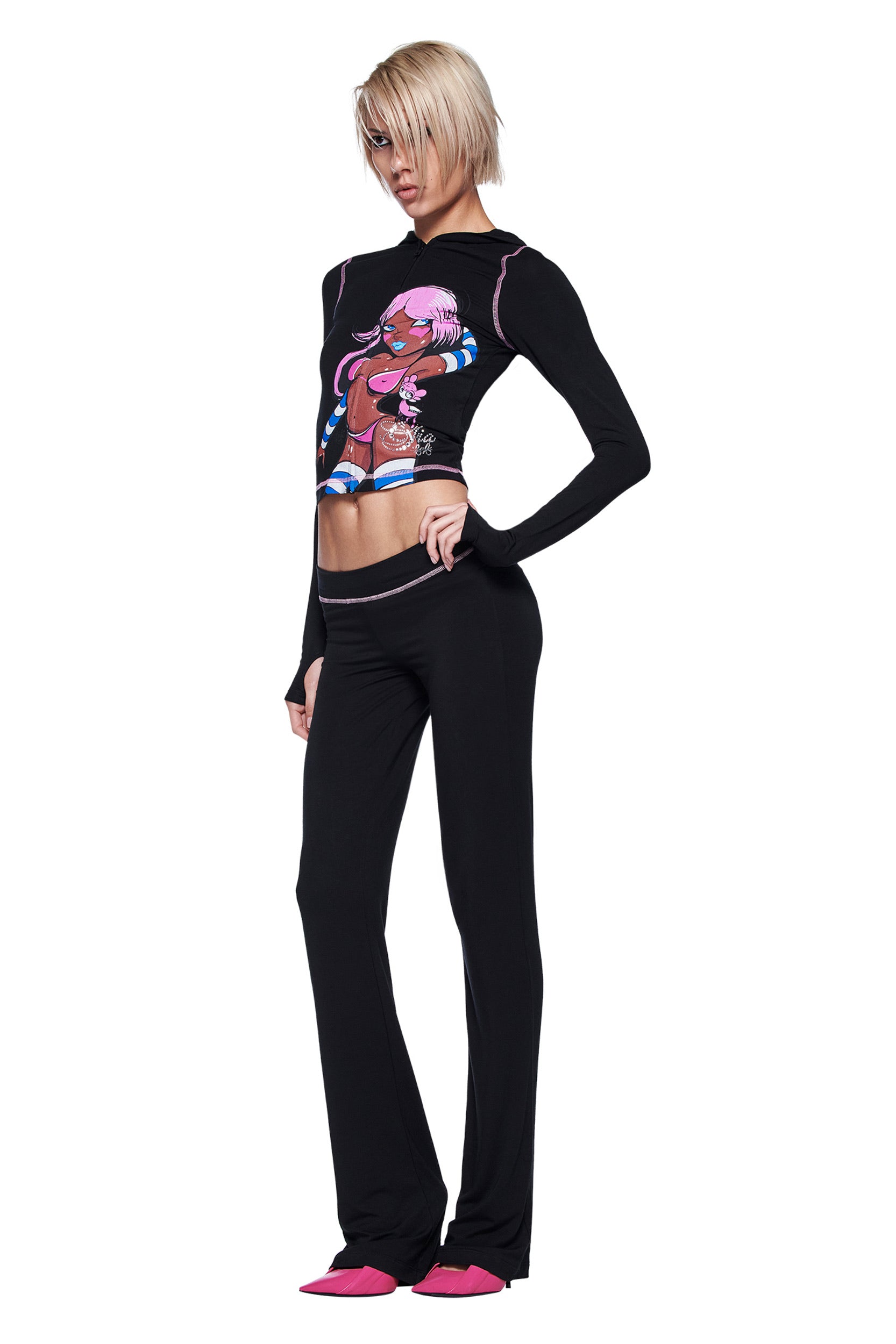 A person with short blonde hair poses against a white background, wearing a black long-sleeve Gia X Fafi top with colorful anime art, I.AM.GIA's FAFI BLARE PANT in black, and pink pointed-toe shoes.