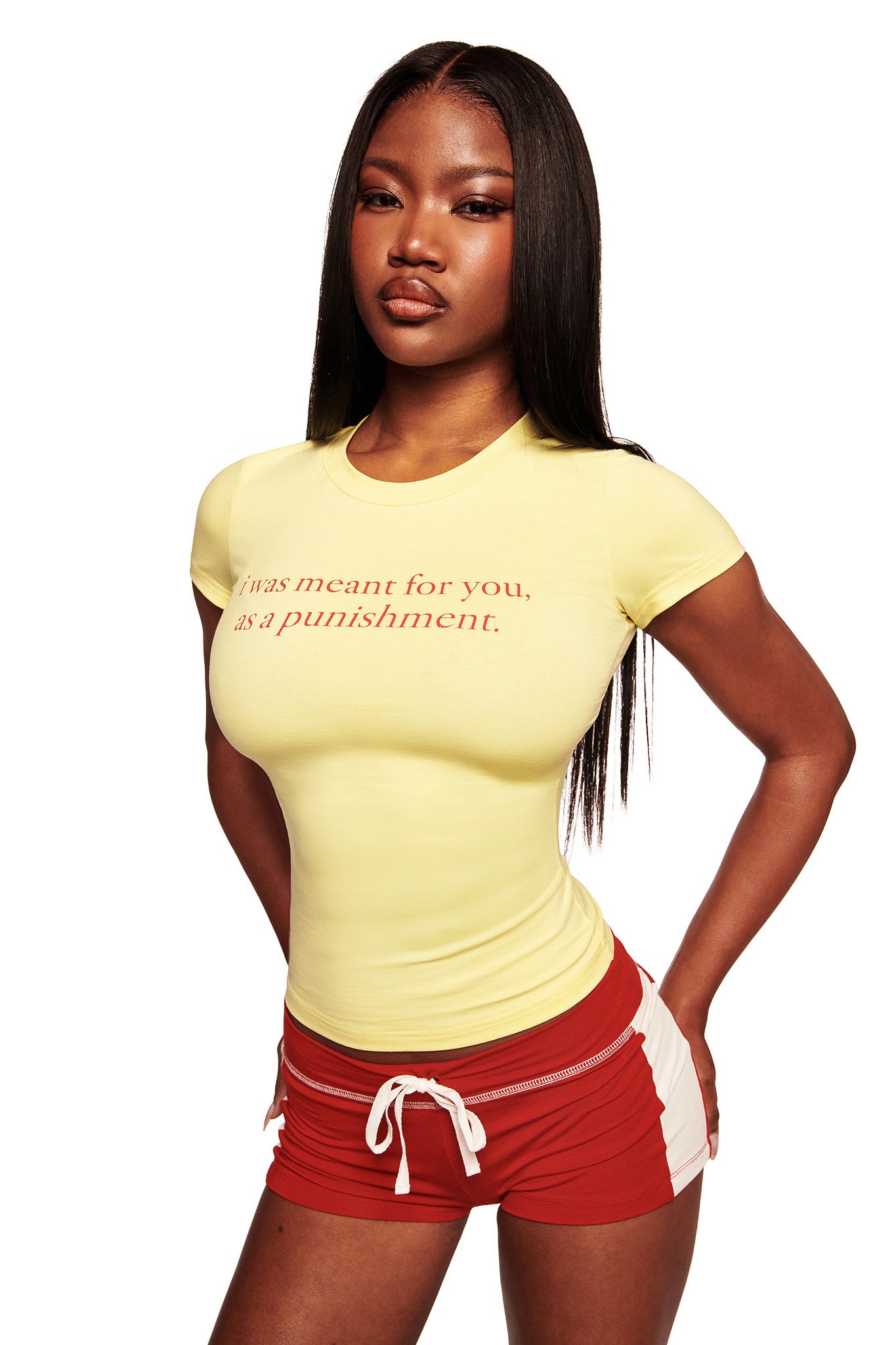 A woman with long, straight hair stands against a white background, wearing the I.AM.GIA POLINA TEE - PUNISHMENT, a fitted yellow graphic t-shirt reading "I was meant for you, as a punishment," and red athletic shorts with white trim.