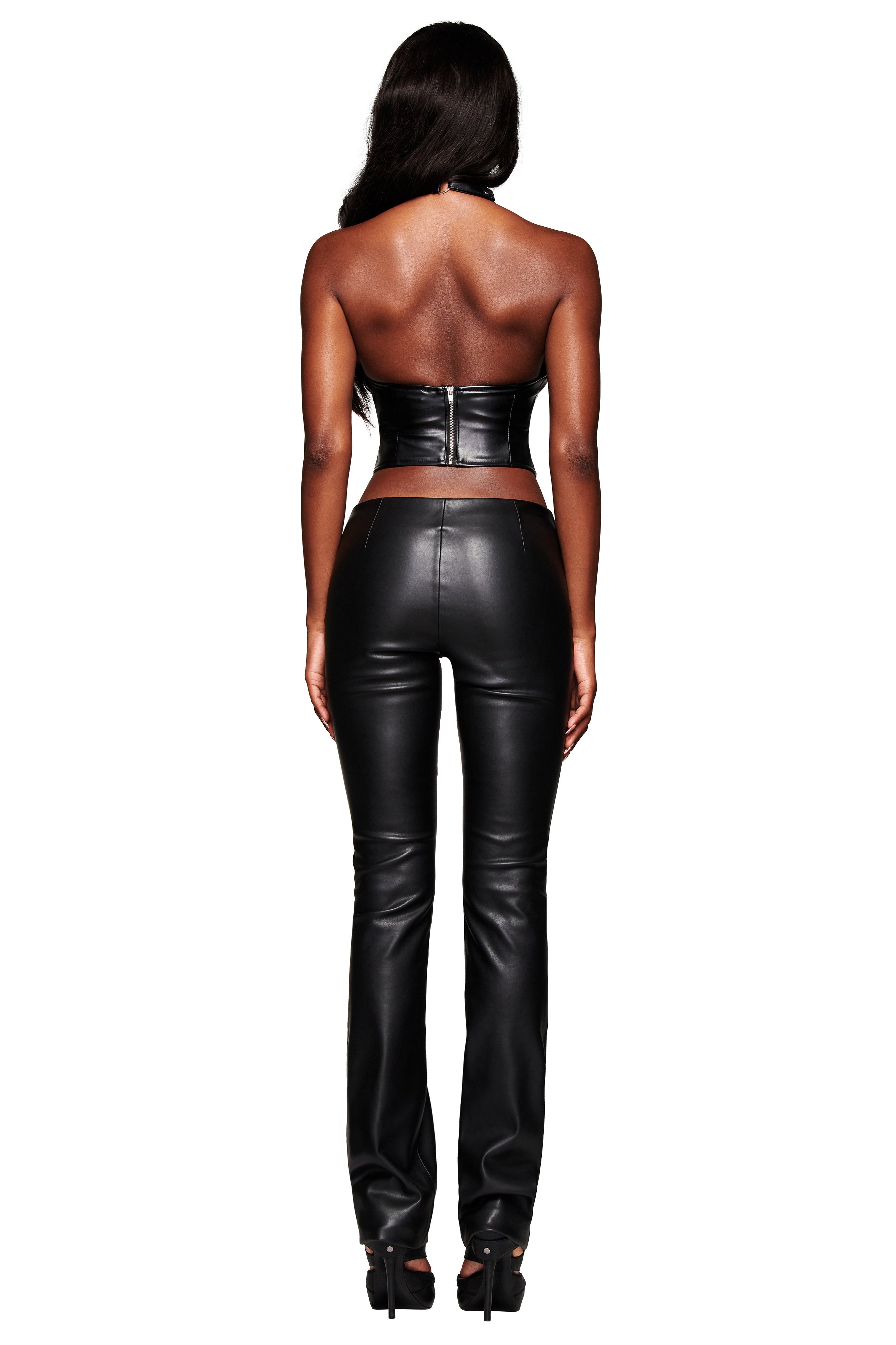 A woman faces away, wearing the I.AM.GIA KENDRIX PANT - BLACK with a straight leg silhouette, paired with a fitted black strapless leather crop top and black heels against a white background.