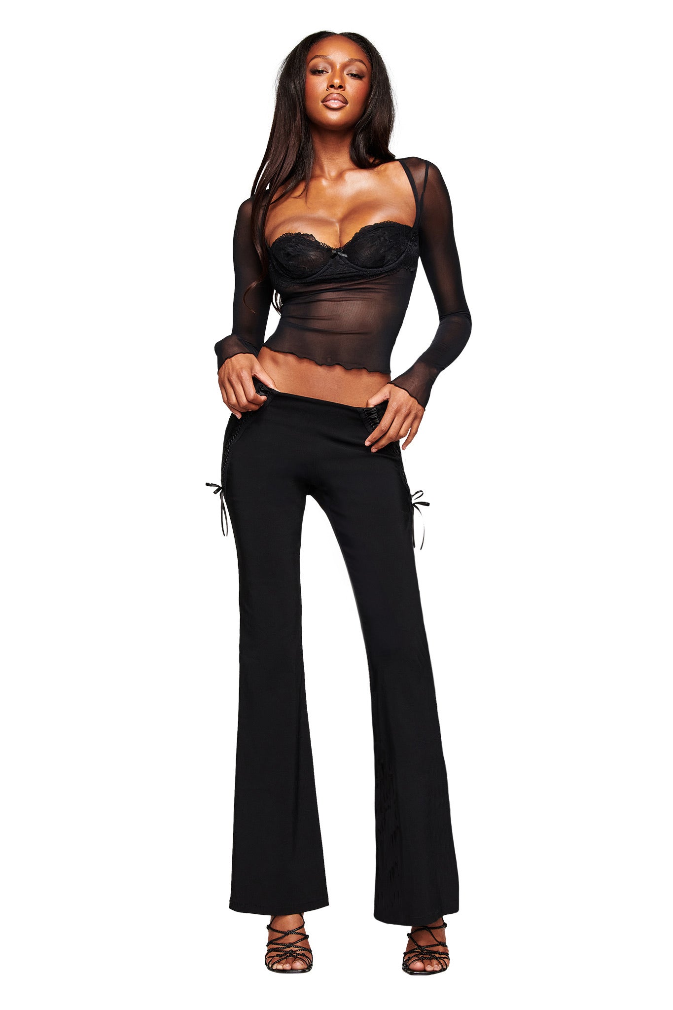 A woman with long dark hair poses against a white backdrop, wearing the I.AM.GIA SILO LONG SLEEVE TOP in black over a bralette, paired with black flared pants and strappy heels.