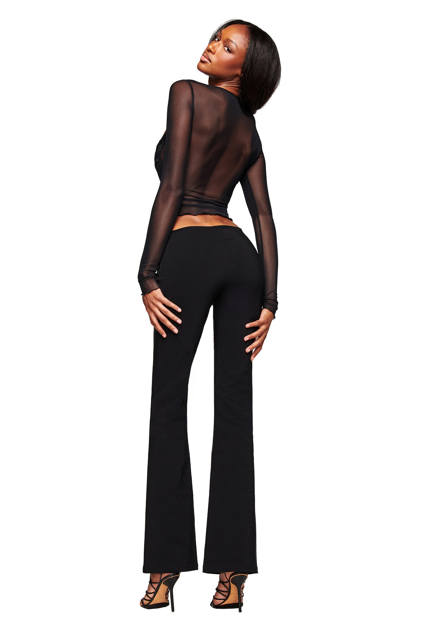 A woman faces backward, glancing over her shoulder and posing against a white background. She wears the I.AM.GIA SILO LONG SLEEVE TOP in black, paired with black flared pants and black high-heeled sandals.