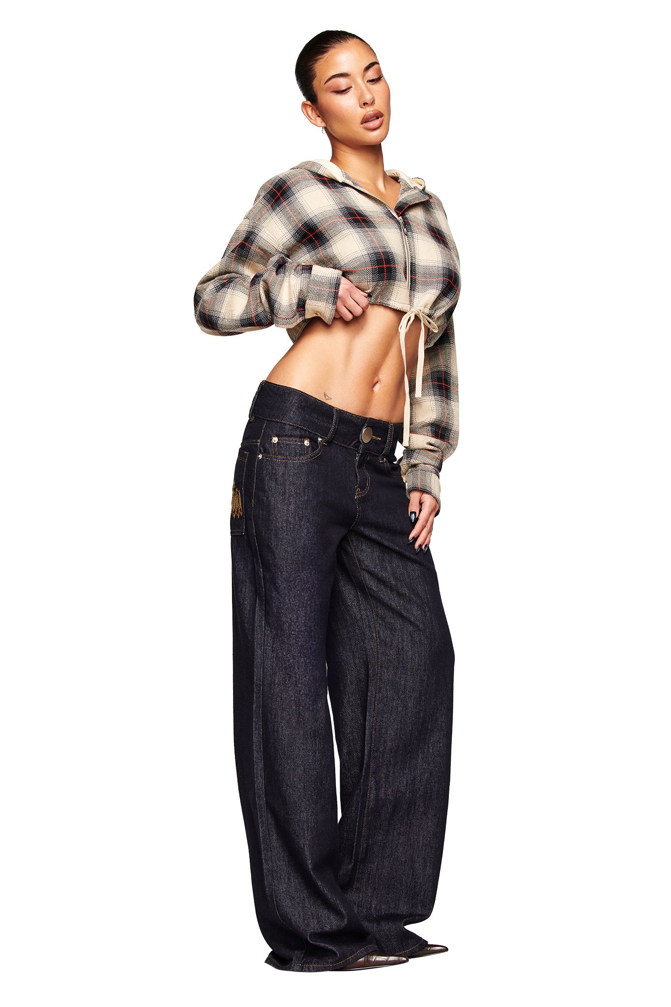 A woman poses in I.AM.GIA’s MOXIE JEAN - INDIGO, styled with a cropped plaid hoodie. She stands against a white background, one hand on her waist and eyes closed, showcasing the low-rise wide-leg jeans and her midriff.