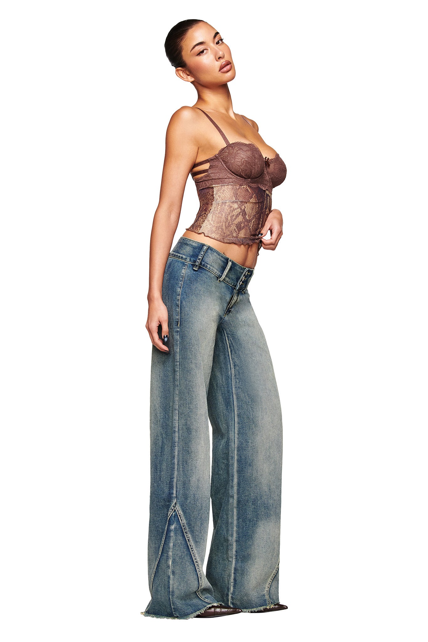 A woman poses against a white background wearing I.AM.GIA’s NYKKO JEAN in blue, featuring visible seams and frayed hems, paired with a sheer brown corset-style top.
