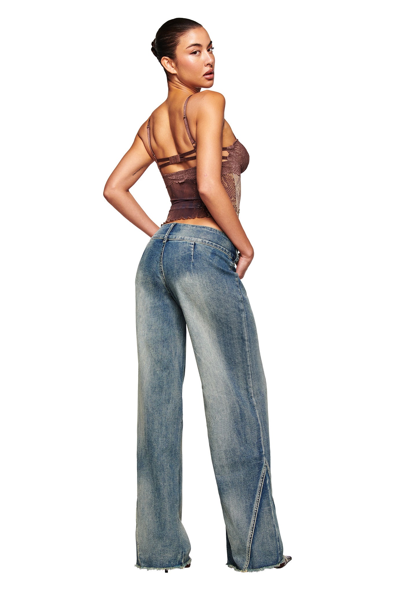 A woman faces away, glancing over her shoulder. She wears a sheer brown camisole and I.AM.GIA NYKKO JEAN - BLUE, featuring low-rise and straight legs with frayed hems. Her hair is in a neat bun against a white background.