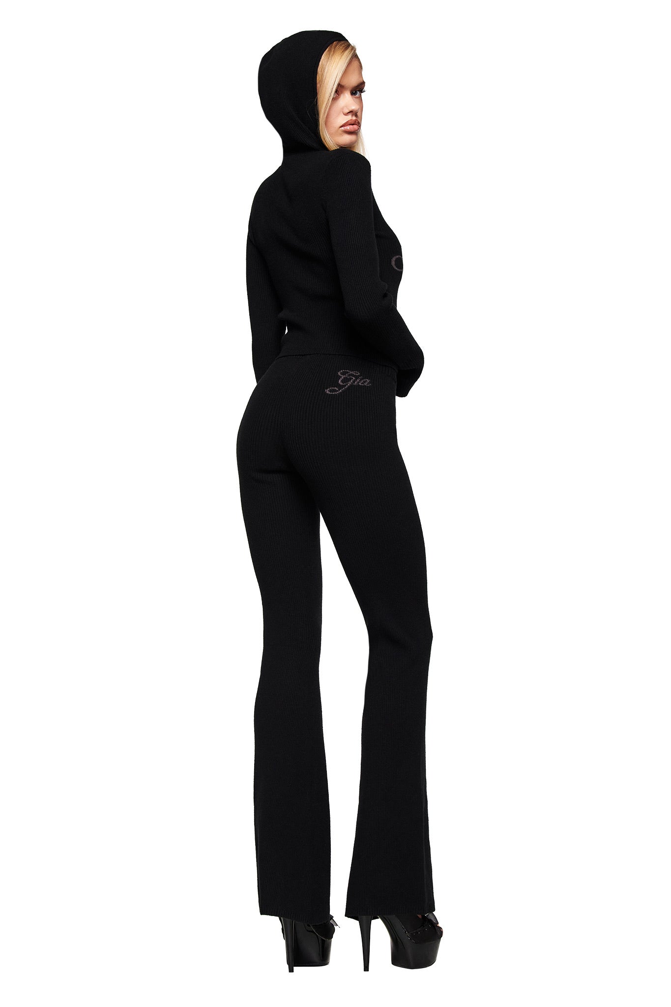 A woman models the I.AM.GIA BLARE KNIT TRACKPANT in black, featuring a mid-rise elasticated waistband and flared legs, paired with a fitted black hooded sweater and high platform heels against a white backdrop.