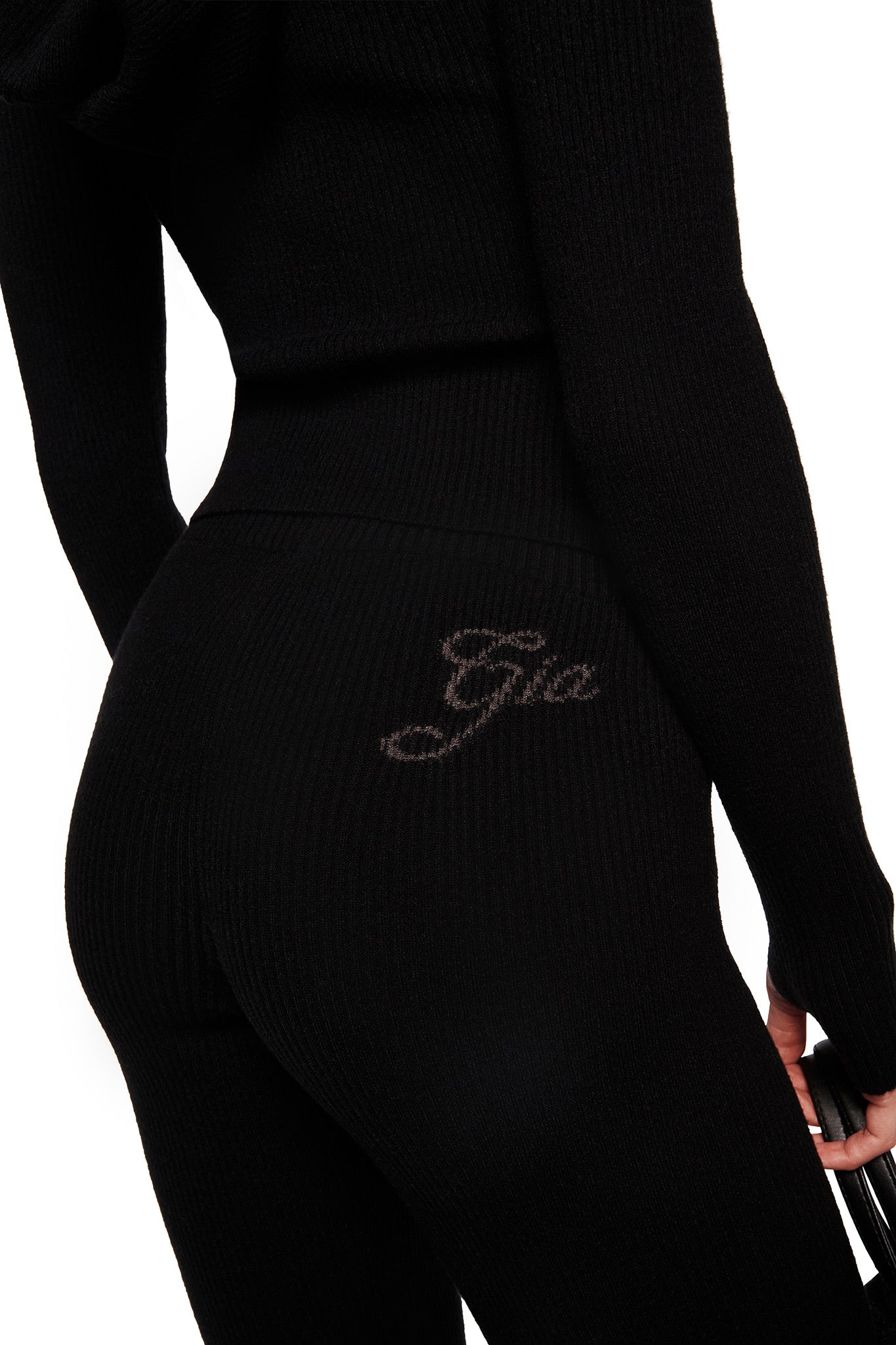Wearing the BLARE KNIT TRACKPANT - BLACK by I.AM.GIA, featuring a ribbed fitted design, mid-rise elastic waistband, and "Gia" cursive embroidery on the upper side, a person holds a dark object at their side with their back and hip visible.