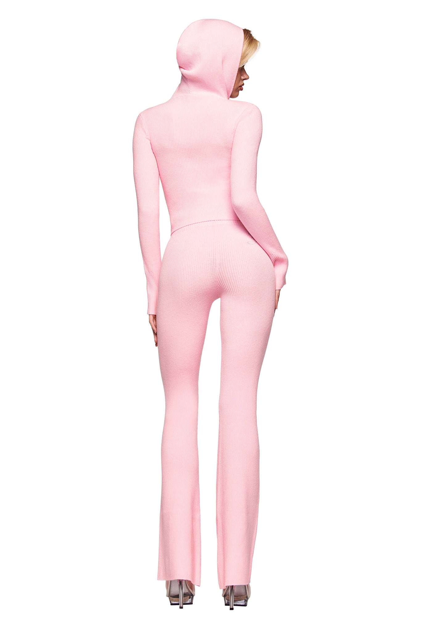 A person stands facing away, wearing the I.AM.GIA BLARE KNIT HOODIE in pink—a bodycon ribbed jumpsuit with long sleeves and flared legs—paired with clear high-heeled shoes.