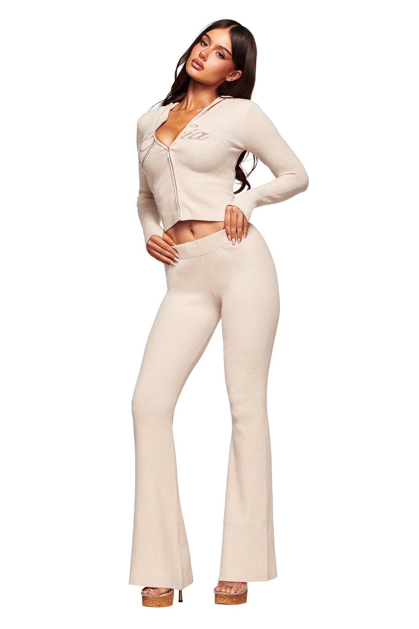A woman models the I.AM.GIA BLARE KNIT HOODIE in cream, a fitted, ribbed knit set featuring a zip-up long sleeve top and matching high-waisted flared pants, styled with platform heels against a plain white background.
