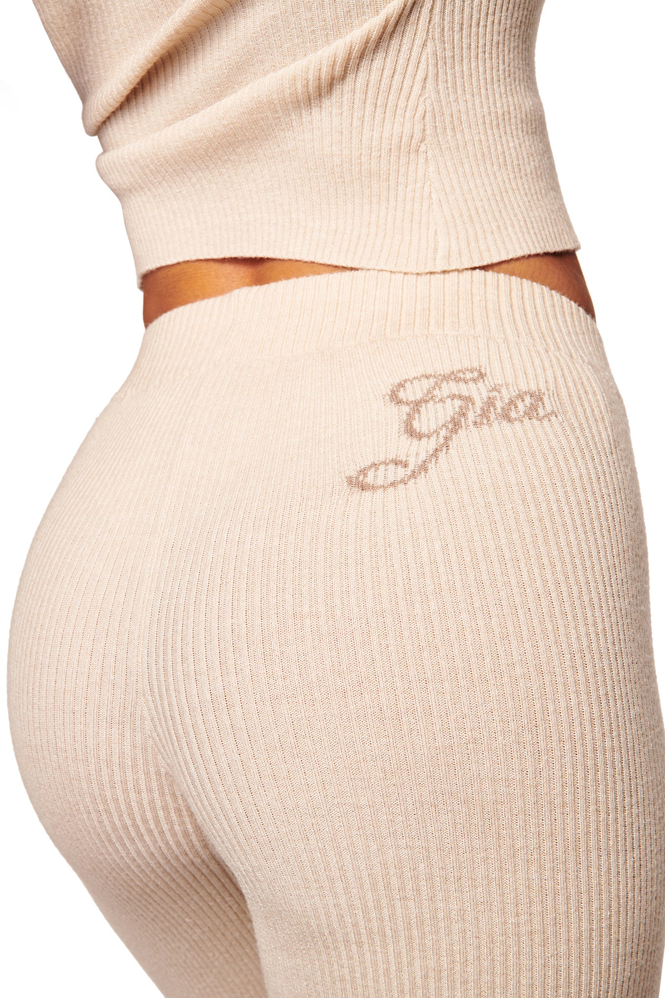 A person wears the I.AM.GIA BLARE KNIT TRACKPANT in cream, featuring a mid-rise elastic waistband with “Gia” script embroidery on the side and a chic flared leg opening.