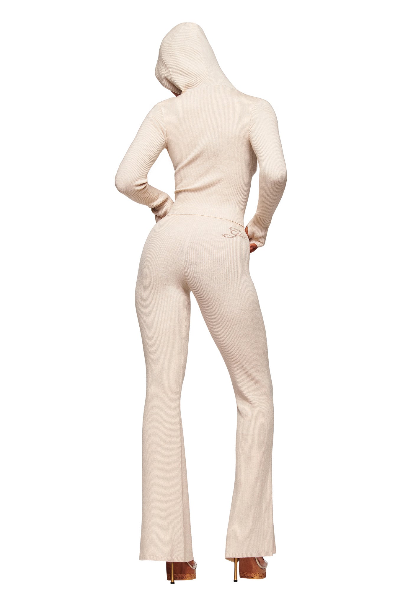 A person stands with their back to the camera, hands on hips, wearing I.AM.GIA’s BLARE KNIT TRACKPANT in Cream with a matching ribbed hoodie, hood up and paired with high-heeled shoes. The trackpant features a mid-rise fit and flared leg openings.