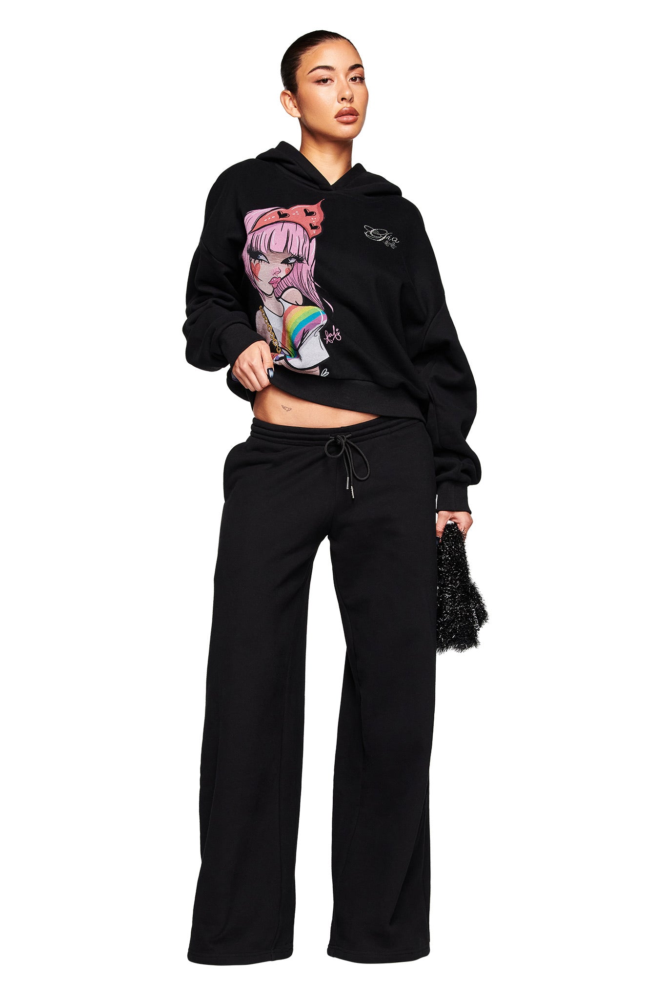 A woman wears the I.AM.GIA FAFI HOODIE - BLACK with anime-style graphics, paired with black wide-leg pants and a small textured black bag, standing against a white background.