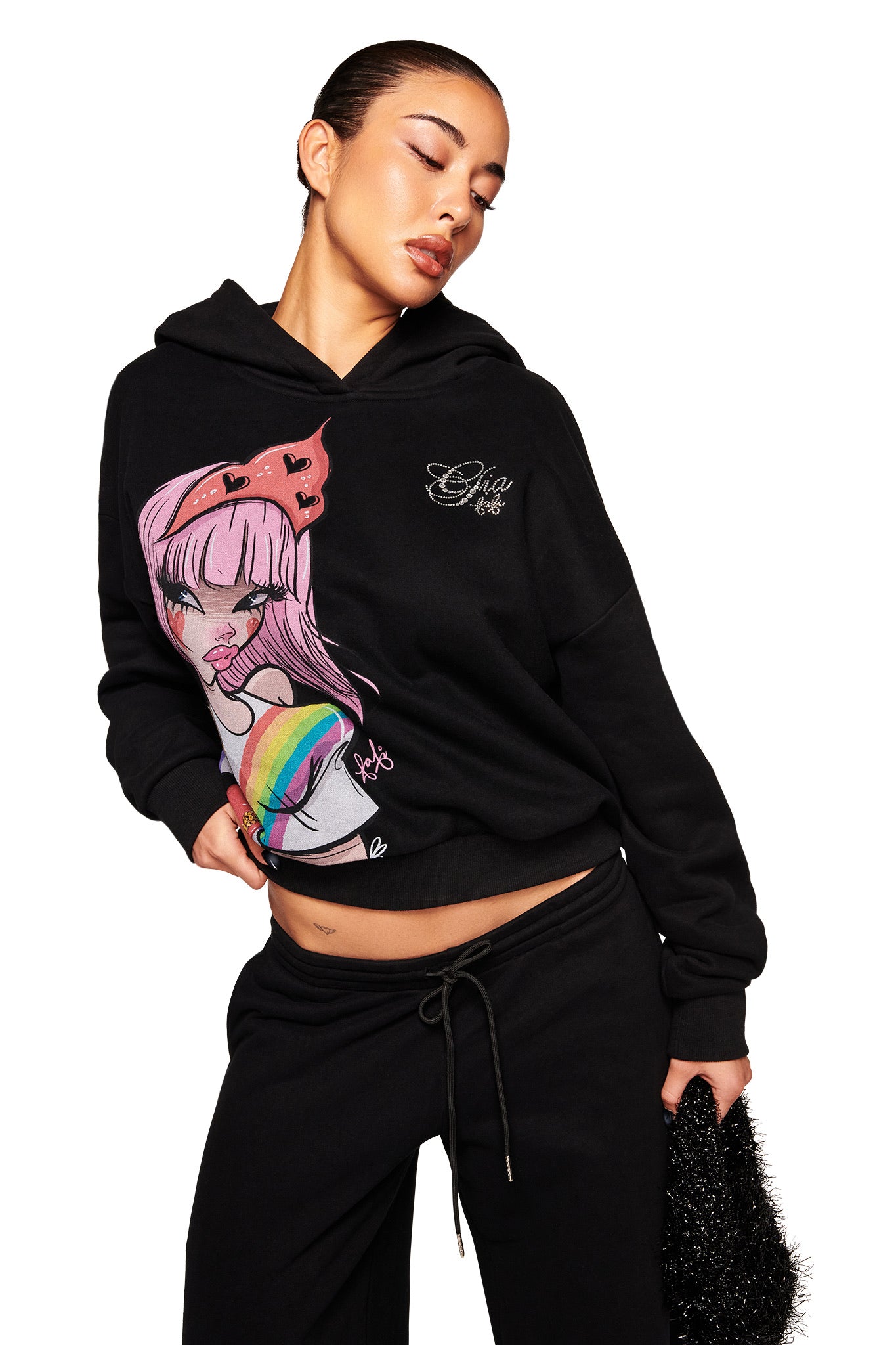 A woman poses against a white background wearing the I.AM.GIA FAFI HOODIE - BLACK, featuring a colorful anime girl graphic, paired with black sweatpants and a fuzzy black bag.