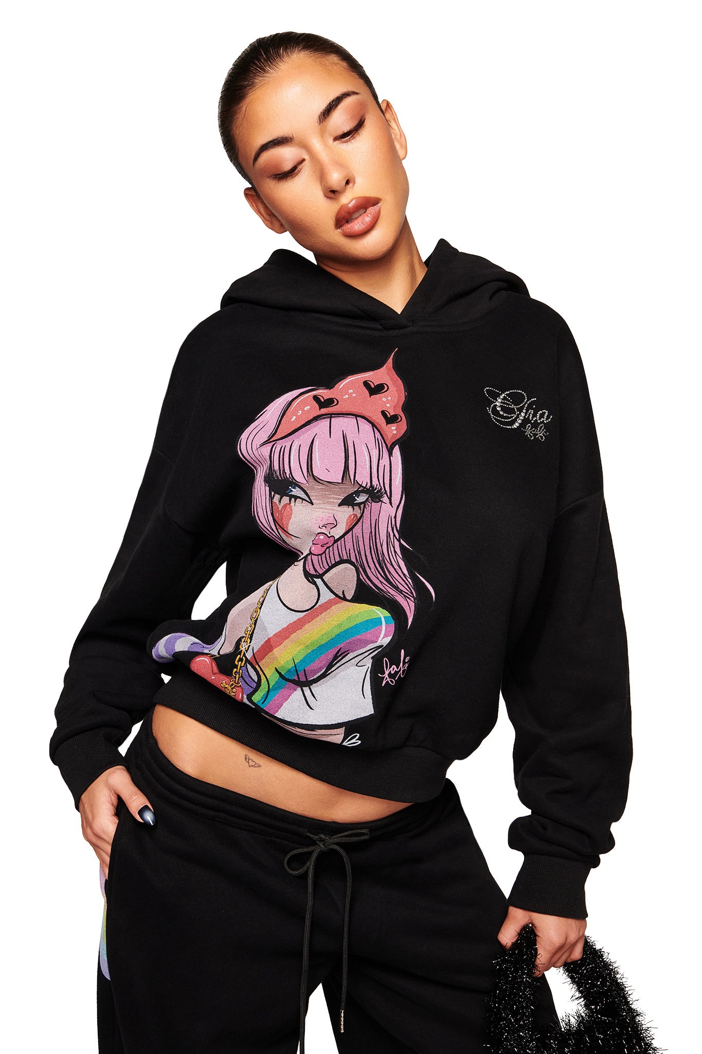 A woman with slicked-back hair models the I.AM.GIA FAFI HOODIE - BLACK, an oversized piece featuring colorful anime-inspired art from the FAFI collaboration. She poses with eyes closed and one hand in her pocket against a white background.