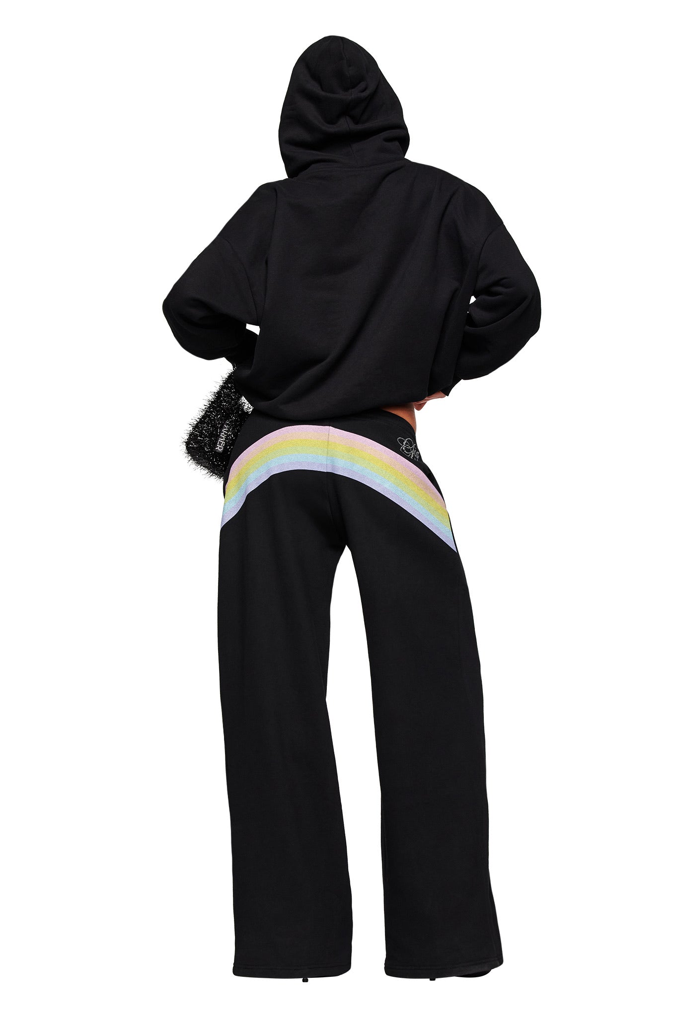 A person wears the I.AM.GIA FAFI TRACKPANT in black, featuring a pastel rainbow stripe across the back, paired with a black hoodie and holding a fuzzy black bag, standing against a plain white background.