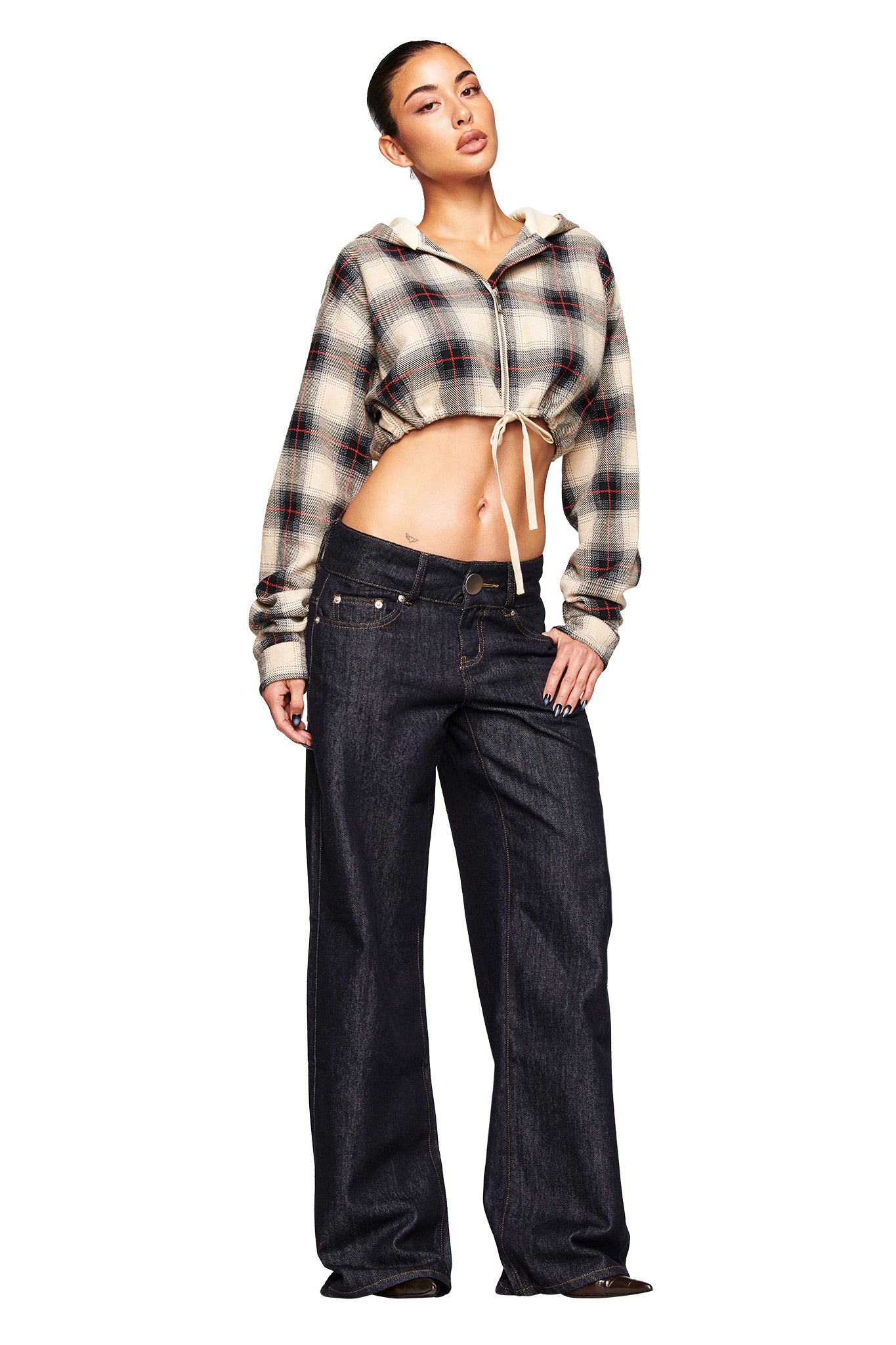 A woman models the I.AM.GIA MOXIE JEAN - INDIGO, pairing the loose-fitting raw denim pants with a cropped plaid hoodie. She poses against a white background, one hand in her pocket, showcasing the jeans' relaxed style.