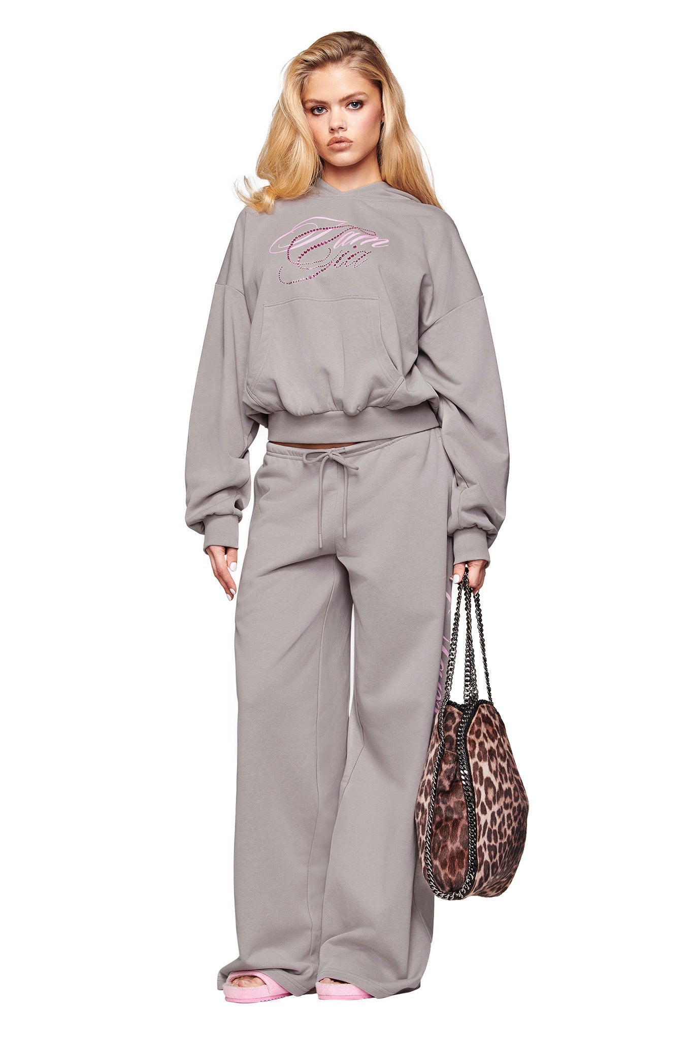 A woman faces forward in I.AM.GIA’s KADE PANT - GREY, paired with a loose grey sweatshirt. She holds two leopard-print bags, wears light pink slippers, and has long blonde hair.