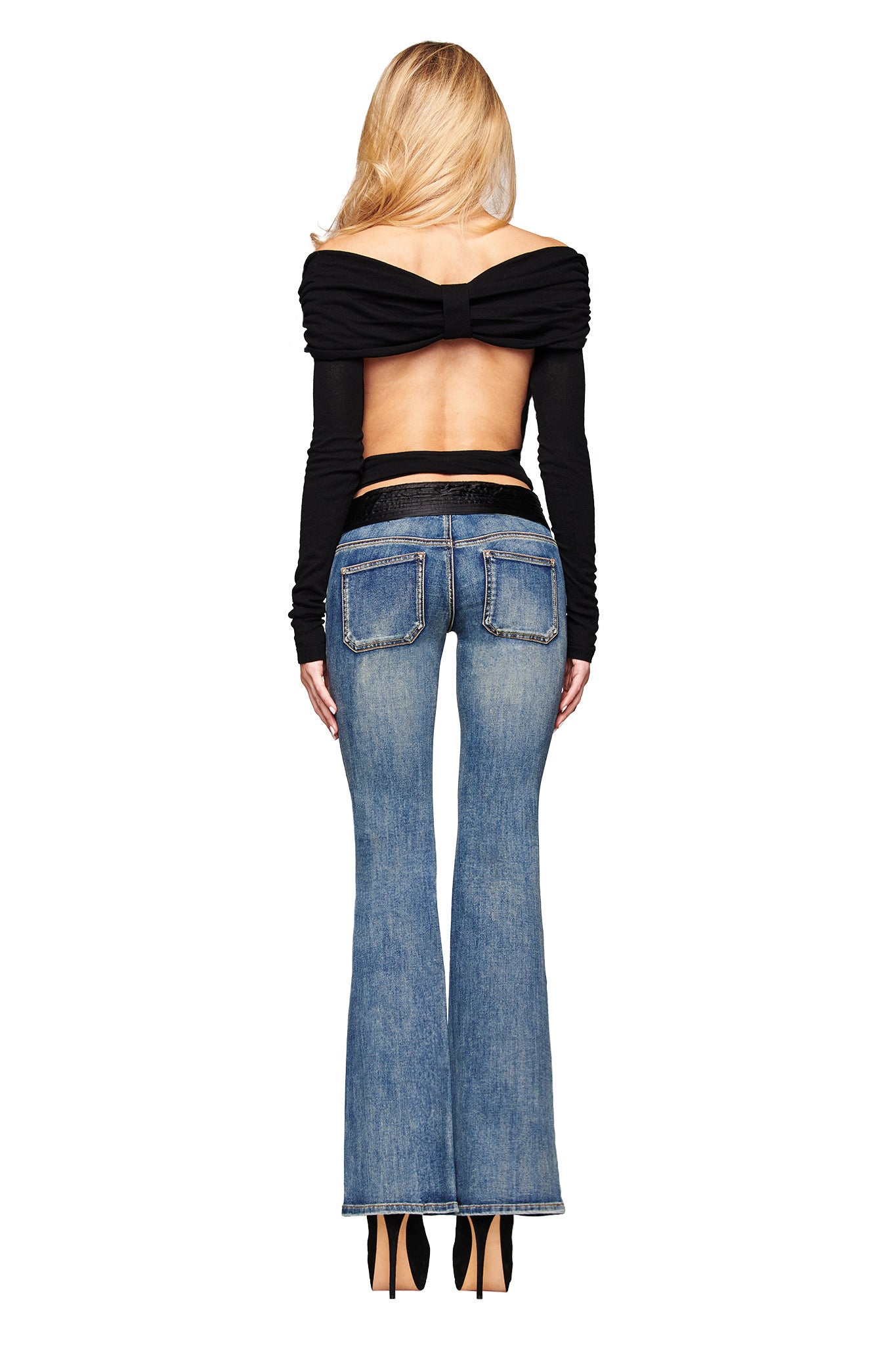 A woman with long blonde hair stands facing away against a white background, wearing I.AM.GIA’s KESHI JEAN in blue denim wash with a flared leg and black satin waistband, paired with an off-the-shoulder open-back black top and high heels.