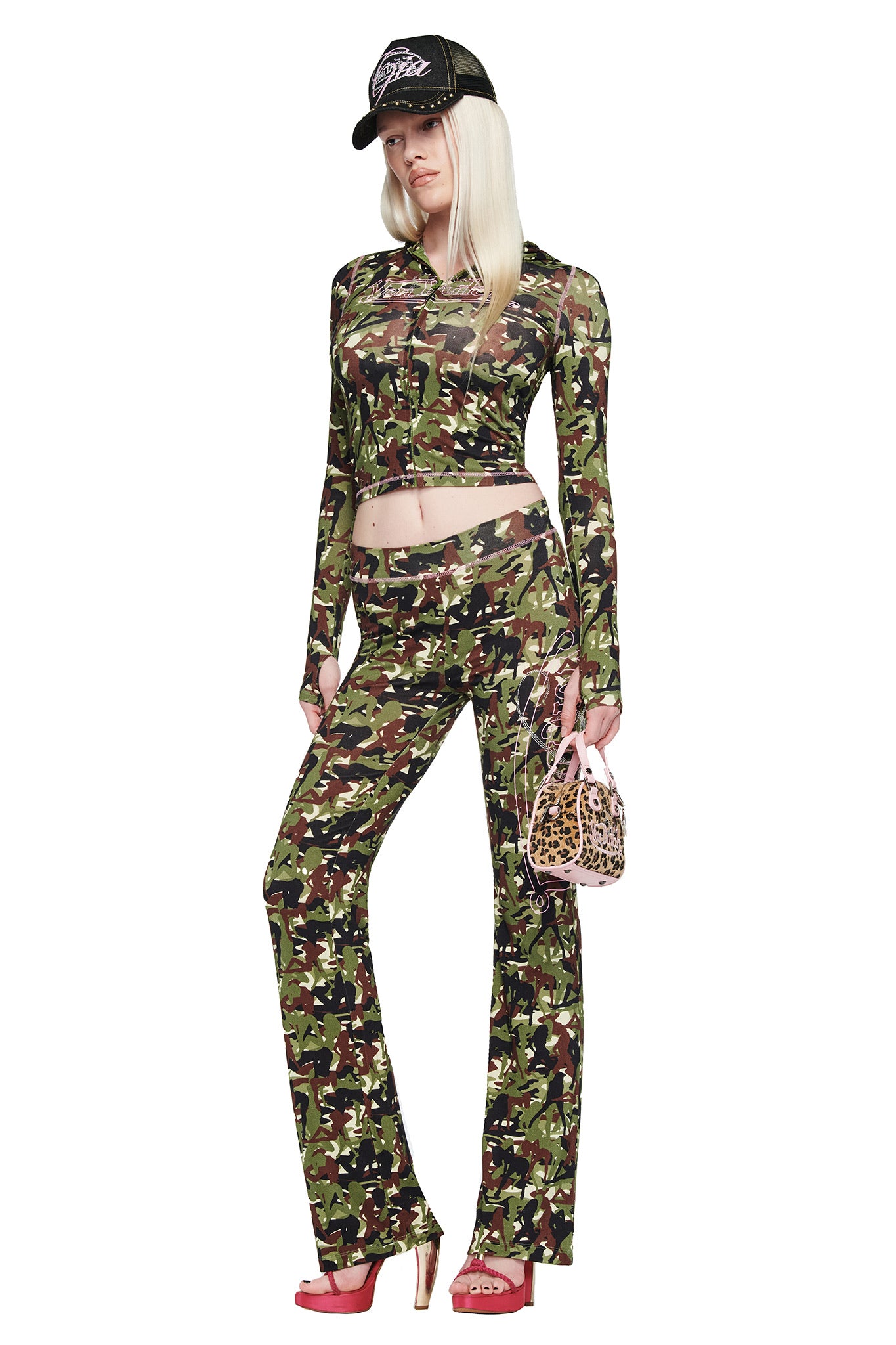 A person stands against a white background wearing the I.AM.GIA X VON DUTCH BLARE TRACKPANT in Gia Girl Camo, a matching camo top, pink high heels, a black cap, and holding a small leopard-print handbag.