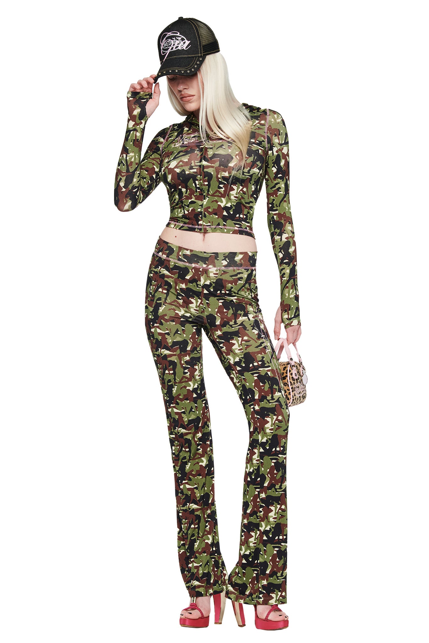A person in a green camo crop top and pants wears the I.AM.GIA X VON DUTCH Trucker Hat in indigo denim, holds a small handbag, and stands in red high heels against a white background.