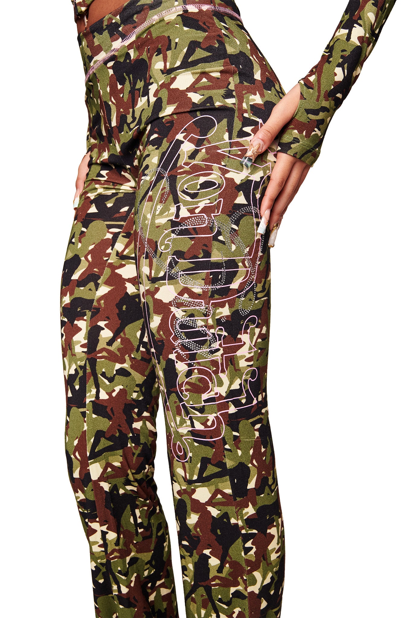 Someone wears the I.AM.GIA X VON DUTCH BLARE TRACKPANT - GIA GIRL CAMO in green and brown camo with white script on the hip, paired with a matching long-sleeve top. The bamboo fabric adds comfort as they rest one hand on their hip.