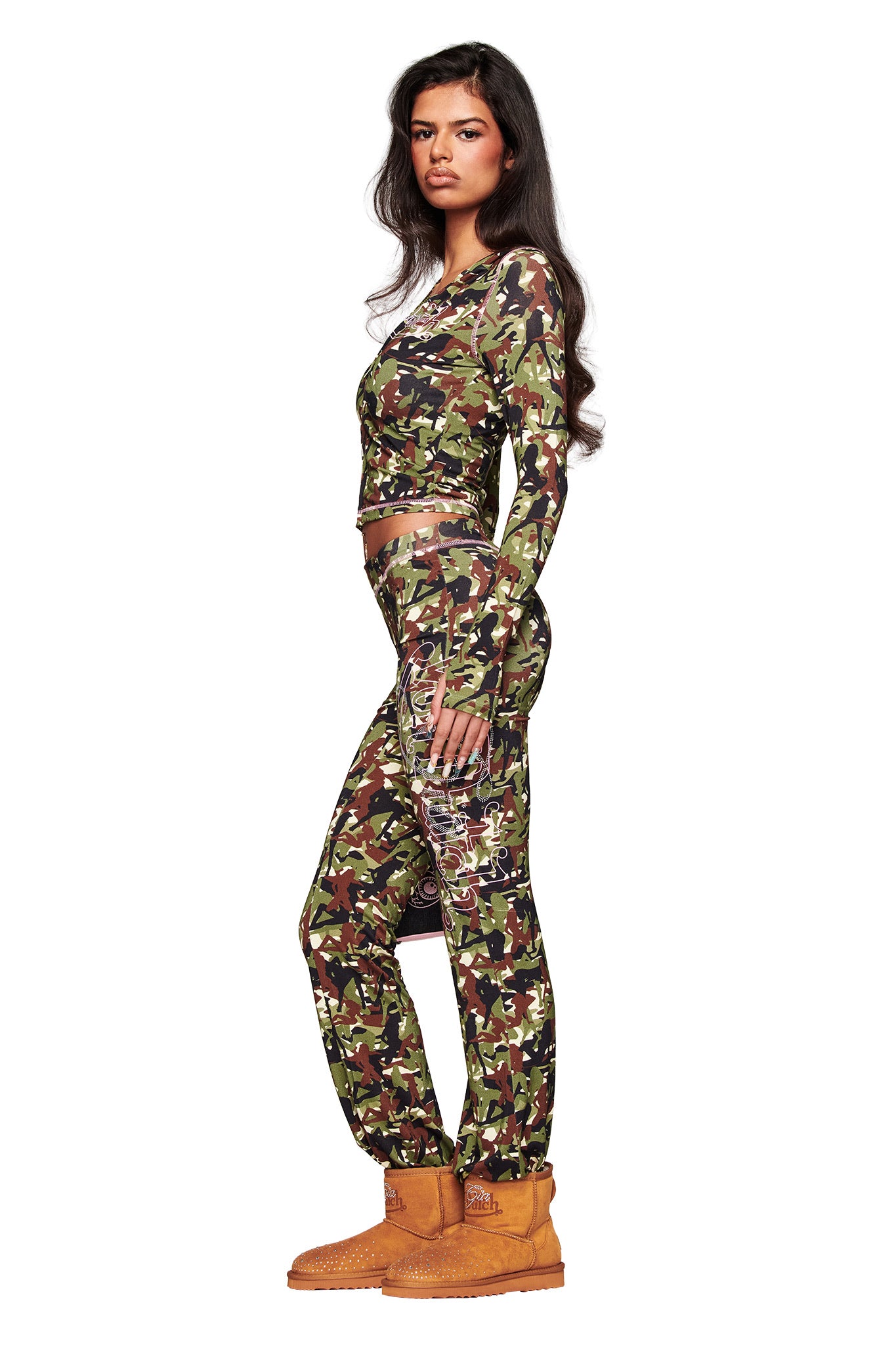 A woman in profile wears the I.AM.GIA X VON DUTCH BLARE TRACKPANT - GIA GIRL CAMO with a fitted camo crop top and tan work boots. She has long, wavy dark hair and a neutral expression against a clean white background.