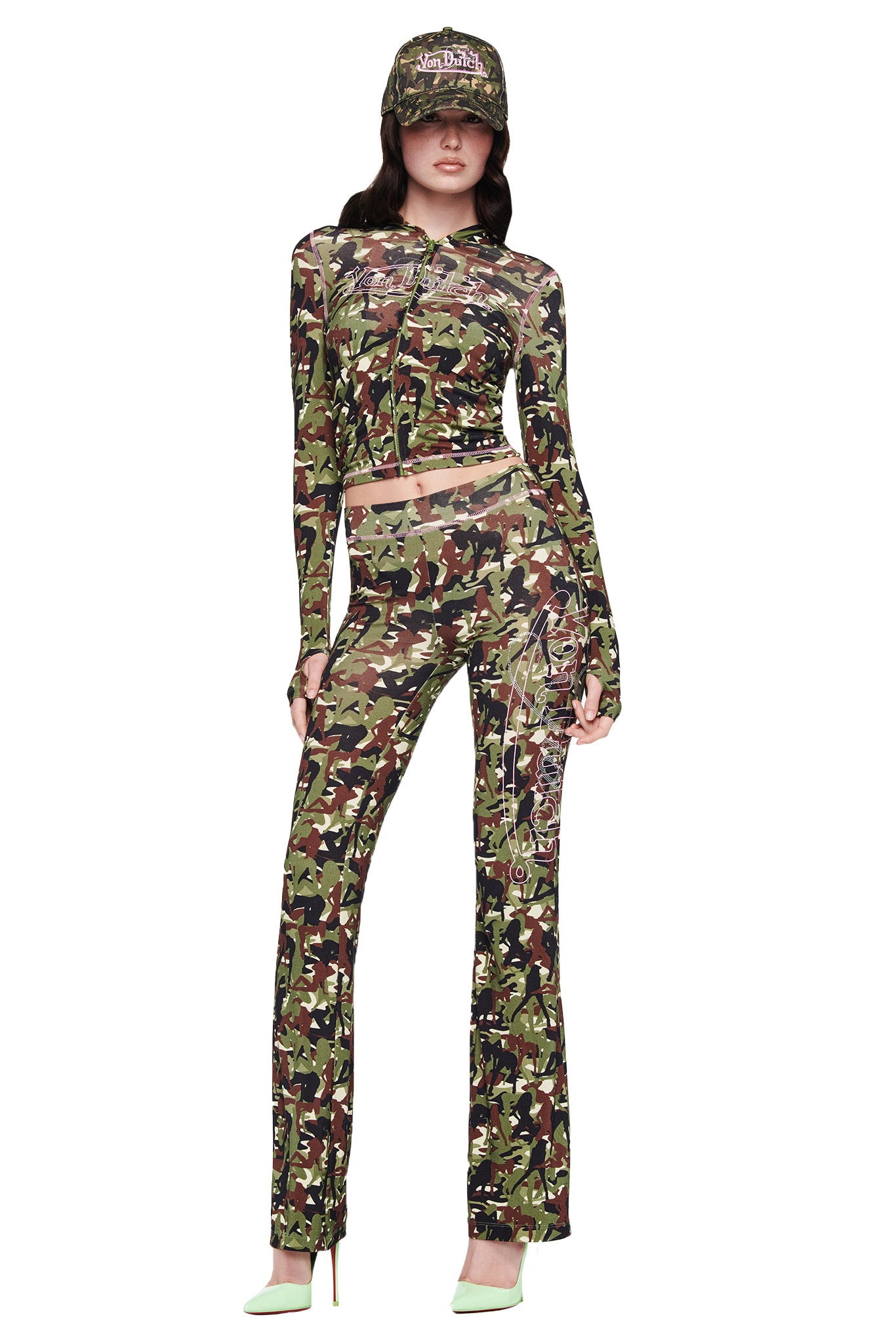 A woman stands facing forward in a green, brown, and black camo top with matching flared pants, pale green heels, and the I.AM.GIA X VON DUTCH TRUCKER HAT - GIA GIRL CAMO by I.AM.GIA. The background is white.