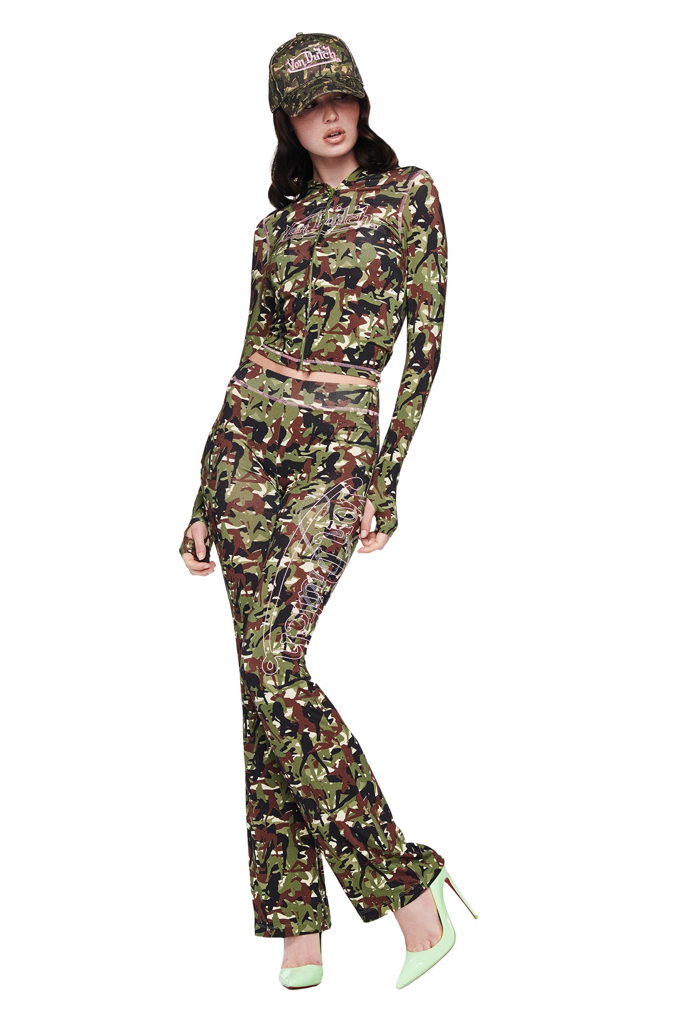 A person models the I.AM.GIA X VON DUTCH BLARE TRACKPANT - GIA GIRL CAMO by I.AM.GIA, paired with a matching fitted long-sleeve camo crop top, camouflage cap, and light green heels in green, brown, and black hues.