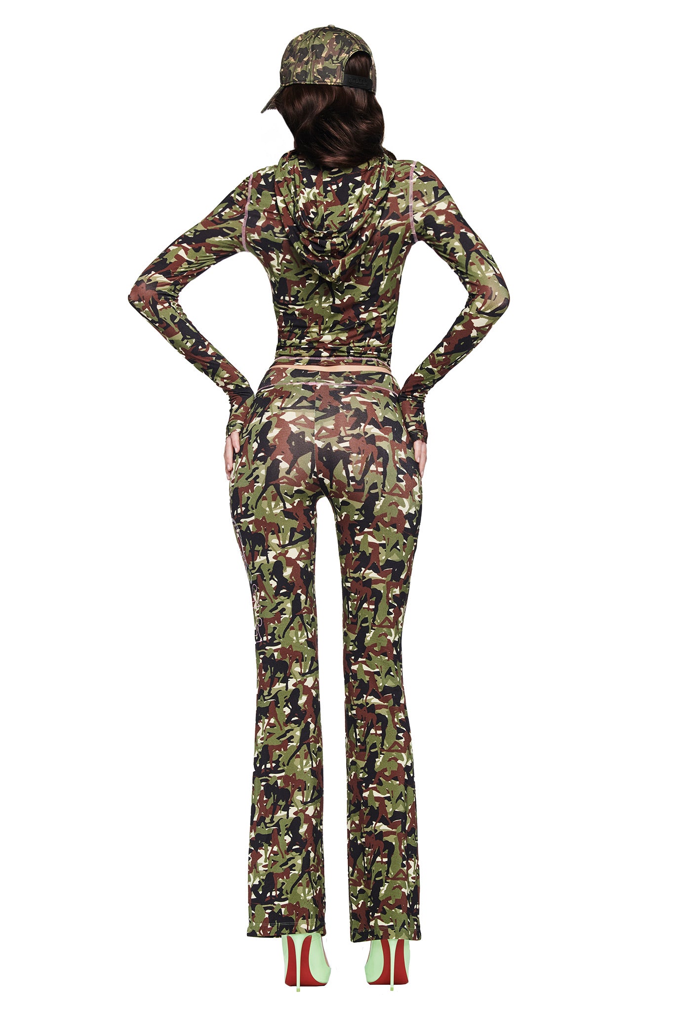 A person faces away in a matching camo jacket, cap, and I.AM.GIA X VON DUTCH BLARE TRACKPANT - GIA GIRL CAMO by I.AM.GIA. Hands on hips, they wear high heels with green soles against a plain white background.