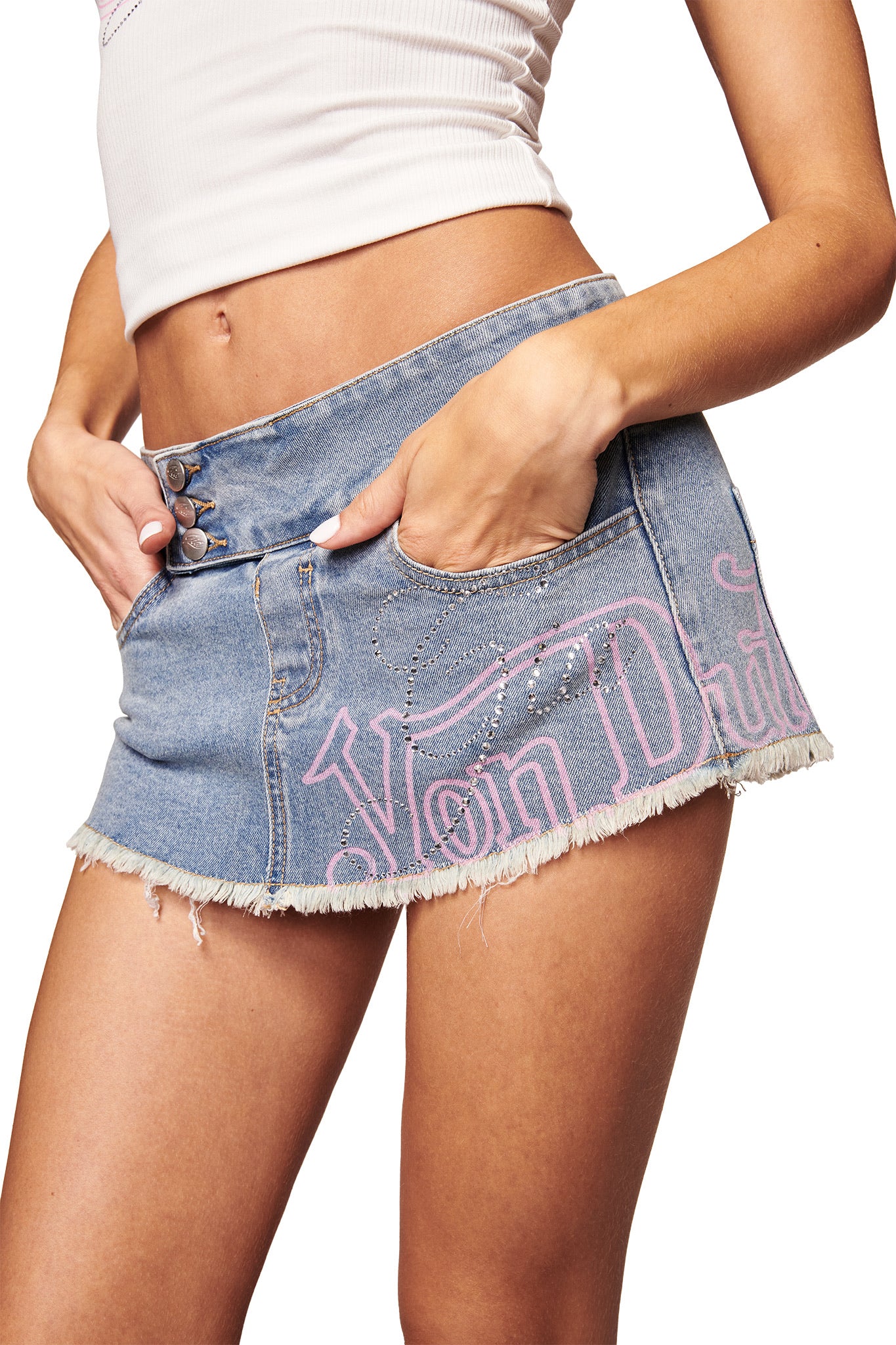 A woman wears a white cropped tank top and the I.AM.GIA X VON DUTCH Syndra Denim Mini Skort in light wash, featuring a low-rise waist, frayed hem, and "Von Dutch" pink logo—a bold statement from the I.AM.GIA collaboration. Hands in her pockets.