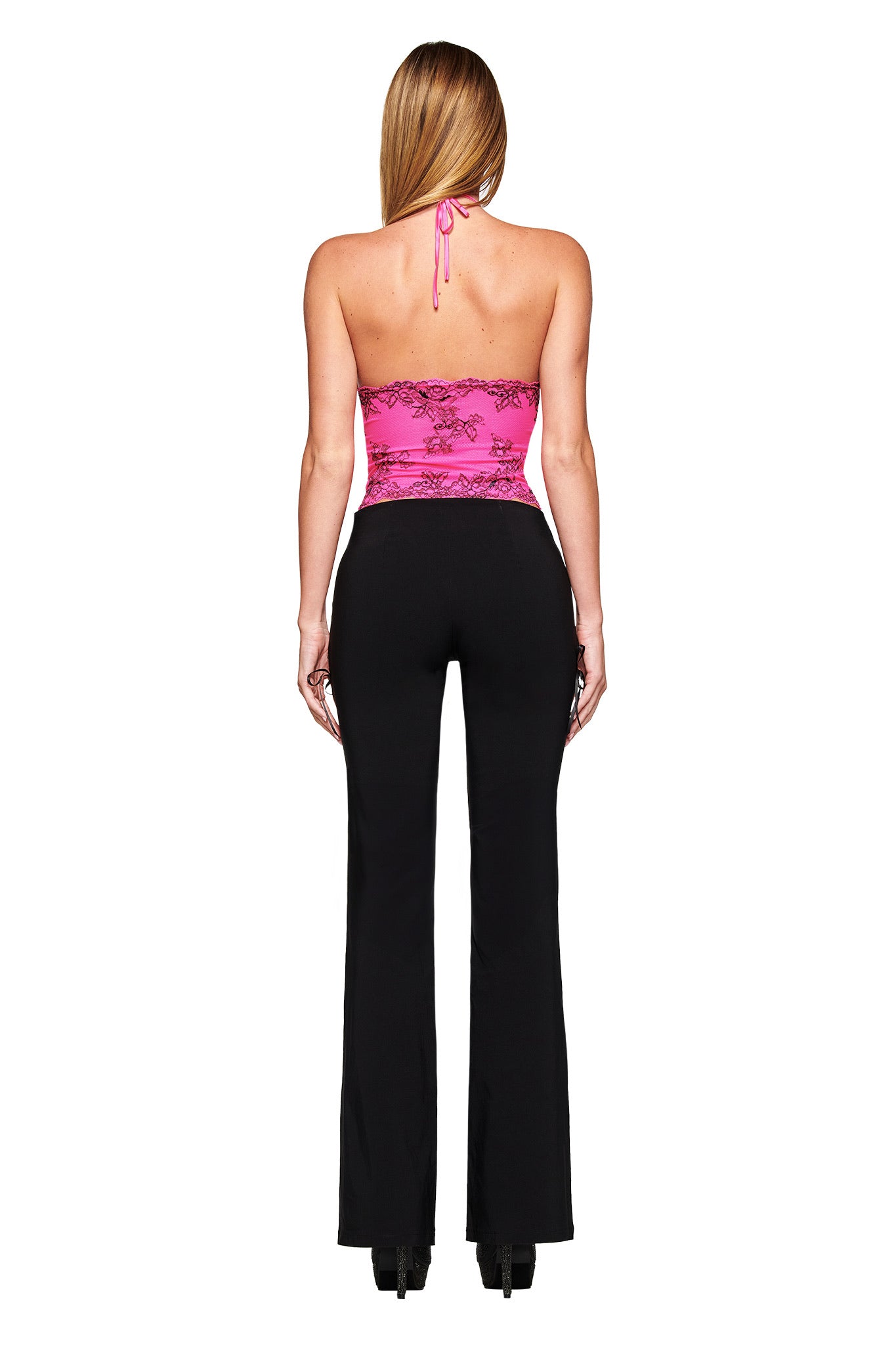 A woman faces away, wearing the I.AM.GIA GEM HALTER TOP in hot pink with black lace and high-waisted black pants. Her long hair is down, and she pairs black shoes with the bodycon look against a white background.