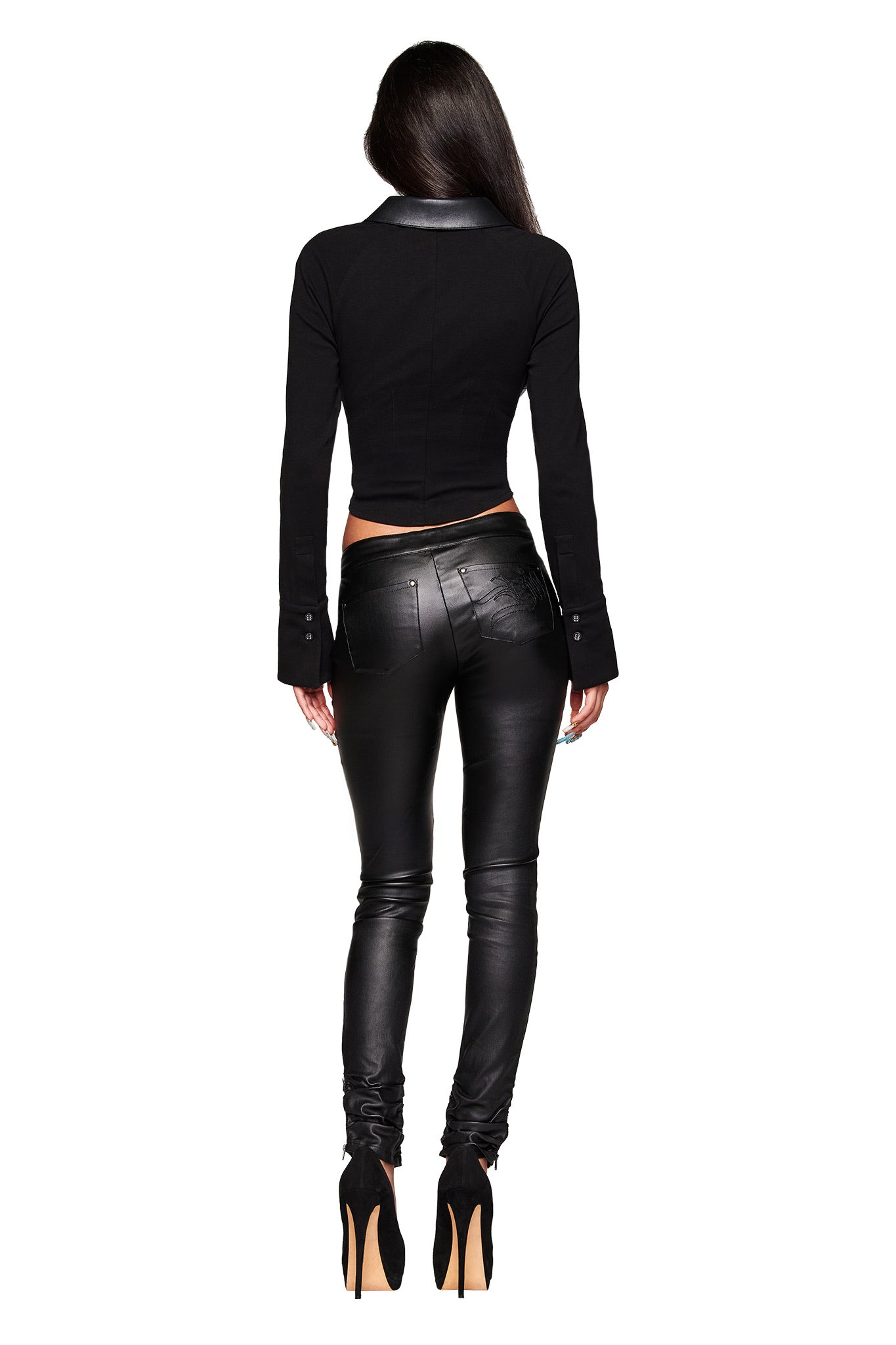 A woman with long dark hair faces away, wearing the I.AM.GIA HELIX TOP in black and shiny bodycon leather pants with high-heeled shoes, set against a white background.