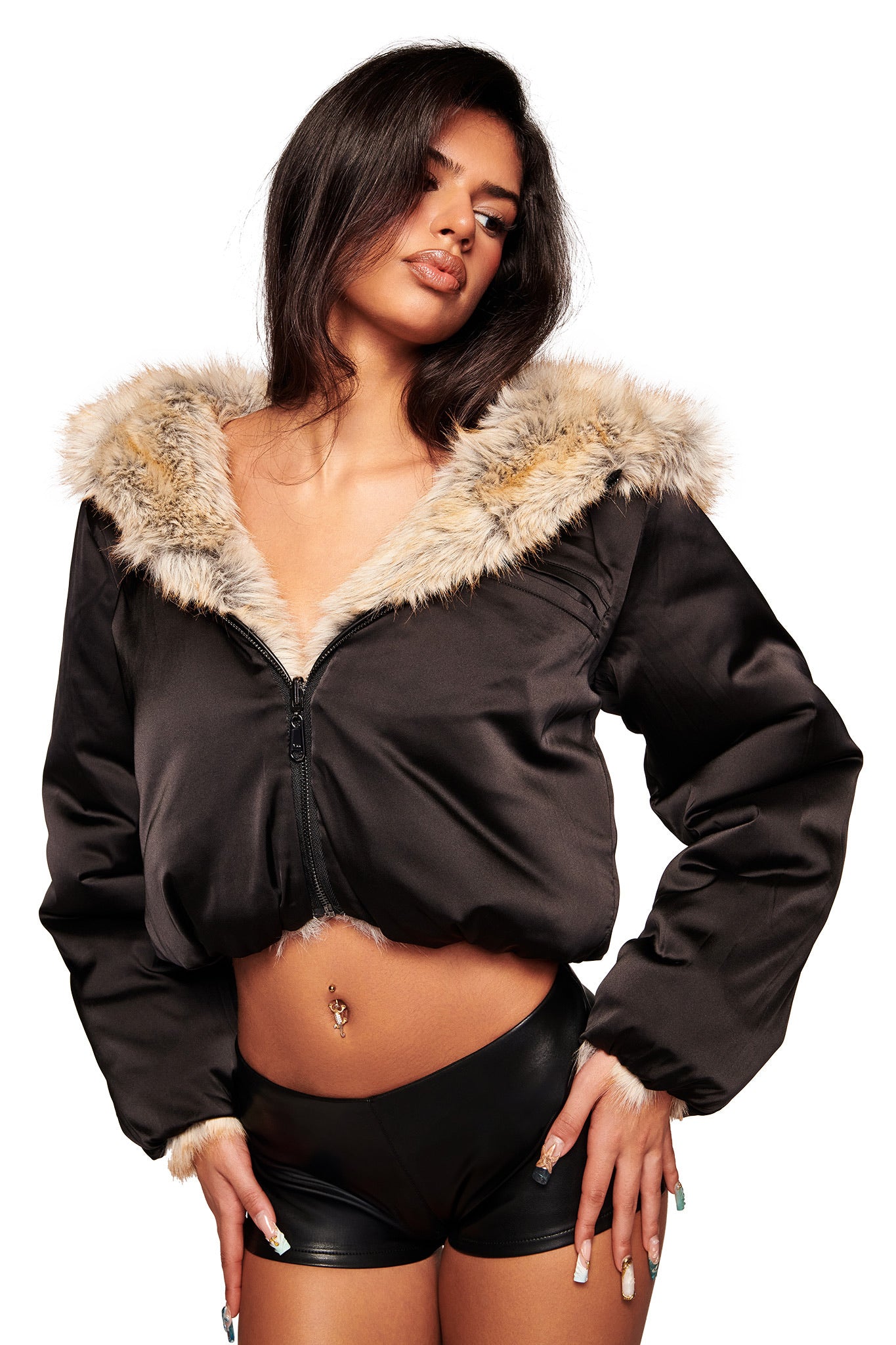 A woman with long dark hair and a belly button piercing poses confidently in the brown EVEREST REVERSIBLE JACKET by I.AM.GIA, featuring a faux fur hood and cuffs, paired with black leather shorts against a white background.