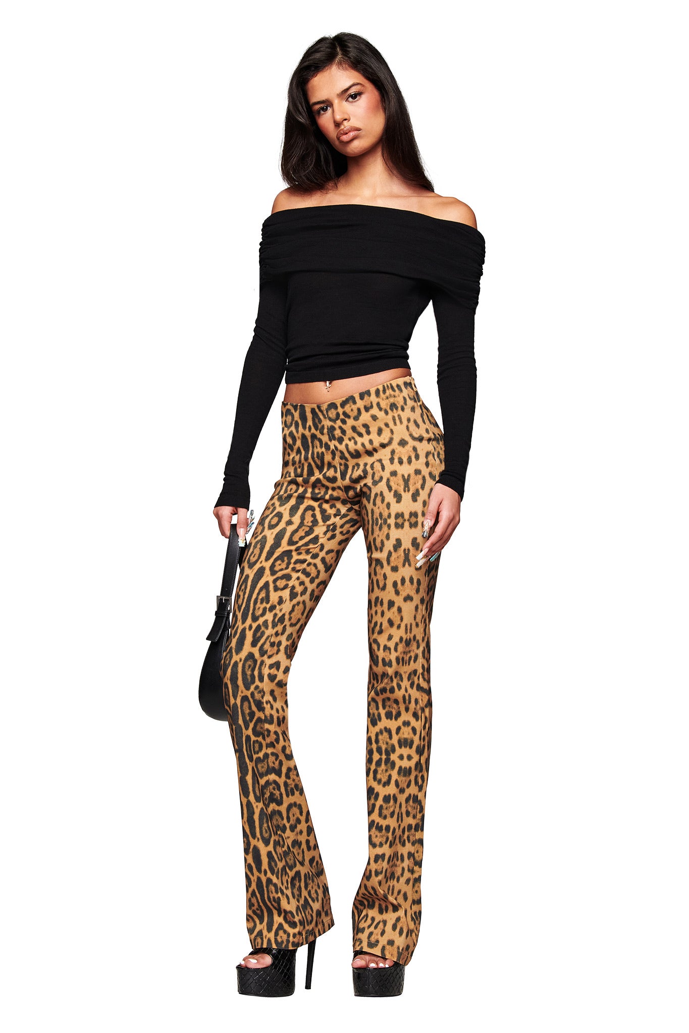 A woman stands confidently in I.AM.GIA UMA PANT - PANTHERA animal print flared pants, a black off-the-shoulder long-sleeve top, black platform heels, and holding a small black handbag against a white background.
