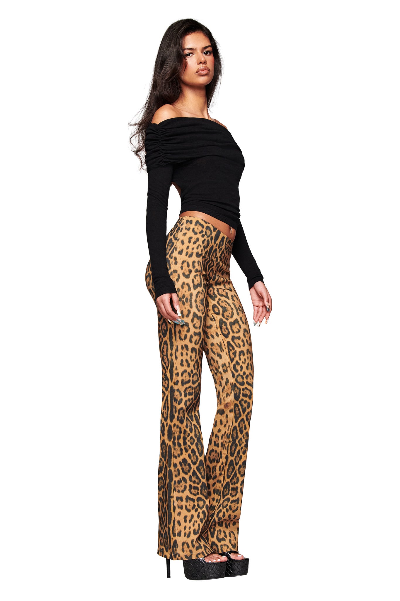 A woman with long dark hair wears the I.AM.GIA UMA PANT - PANTHERA, a pair of high-waisted animal print flared pants, styled with a black off-the-shoulder top and black platform heels against a white background.